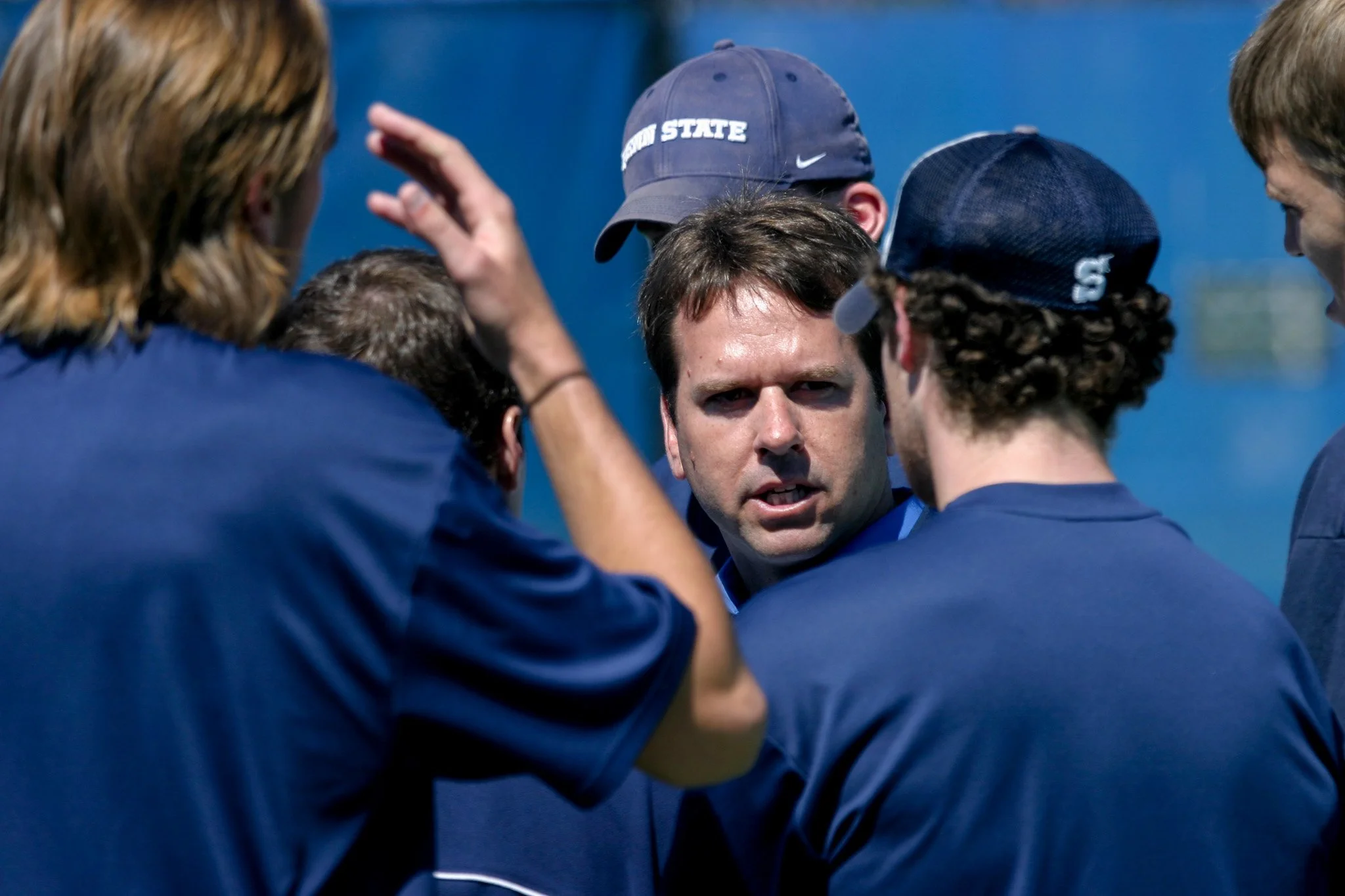 A group of people, likely a sports team or coaching staff, gathered in a huddle outdoors. One person in the center is speaking, with others listening attentively.
