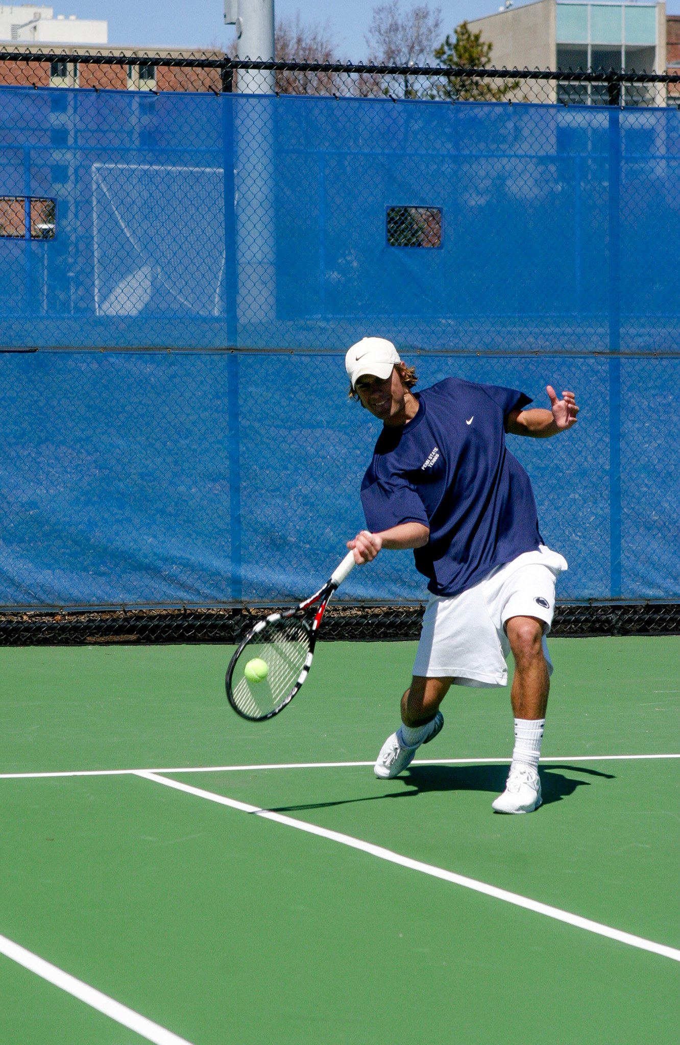 A person playing tennis on a green court, hitting a tennis ball with a racket, wearing a navy blue shirt, white shorts, white shoes, and a white cap, with a blue windscreen and chain-link fence in the background.