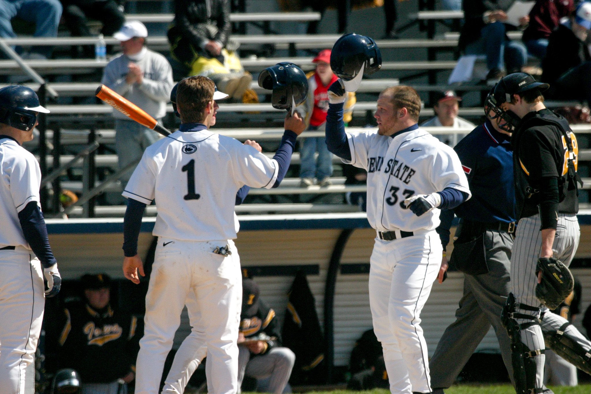 Two baseball players in white jerseys high-five on the field during a game, with spectators sitting on bleachers in the background.