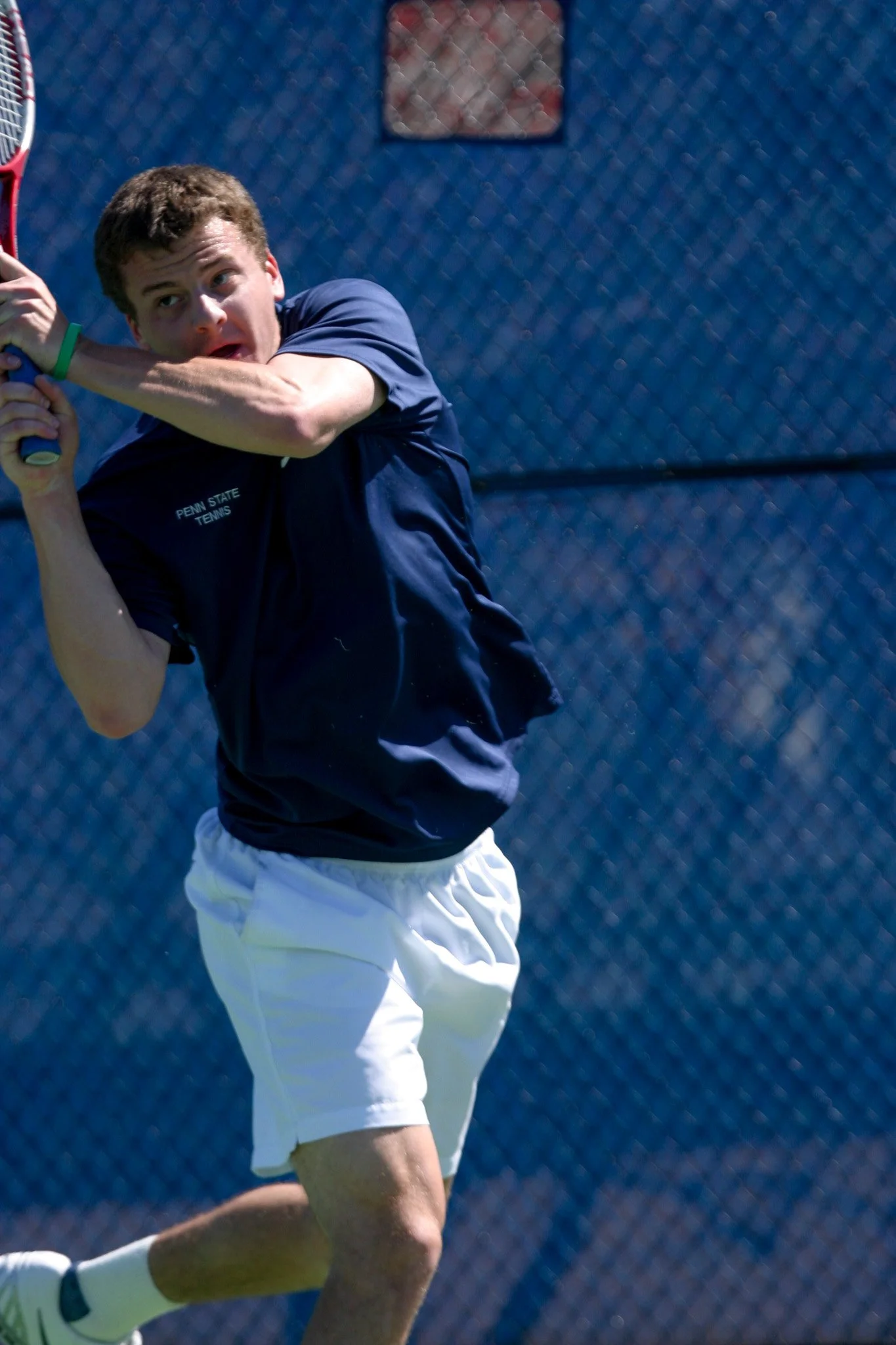 A young man in a navy T-shirt and white shorts playing tennis on an outdoor court, preparing to hit a shot.