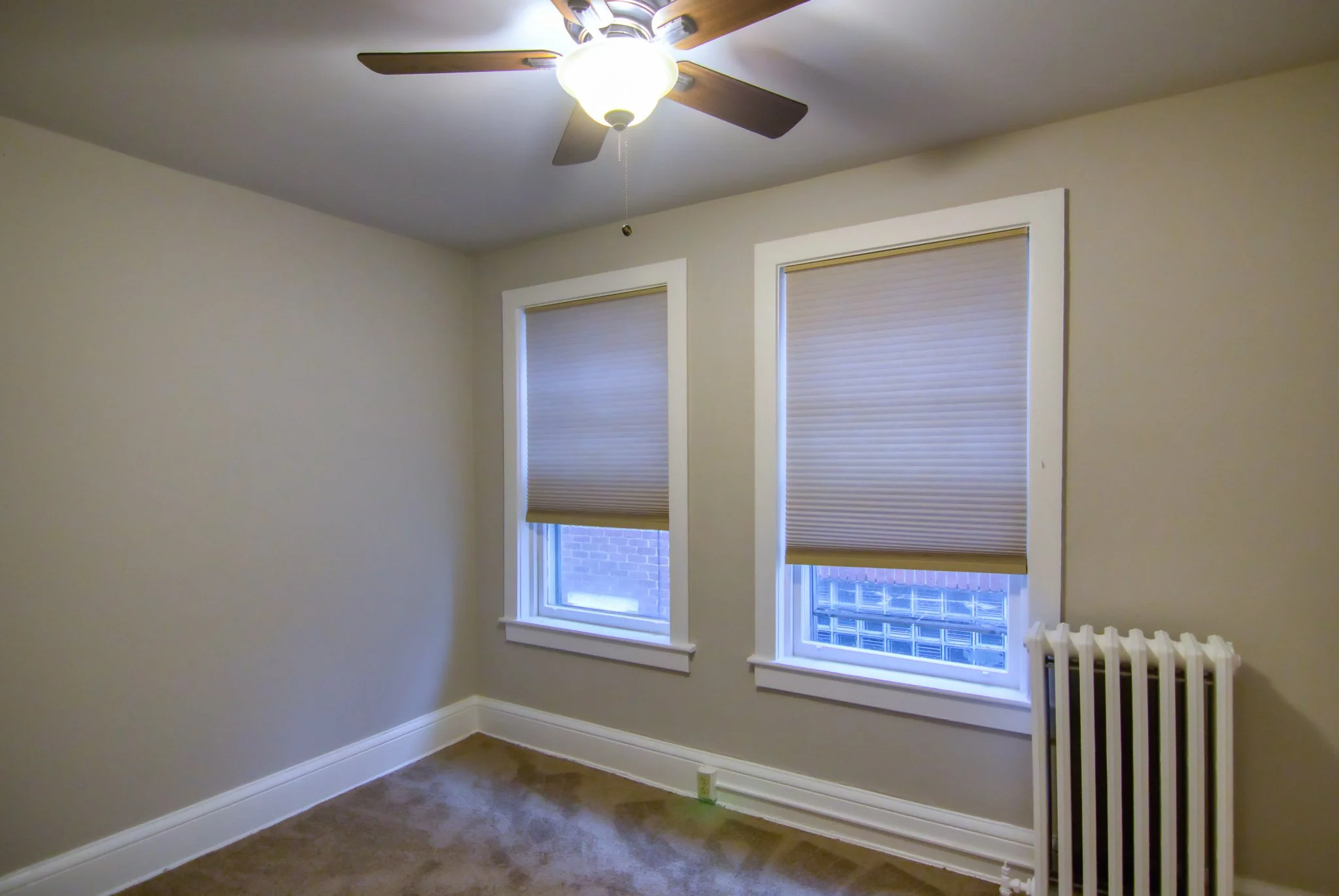 Empty room with beige walls, two windows with beige blinds, a ceiling fan with a light, a radiator, and beige carpet flooring.