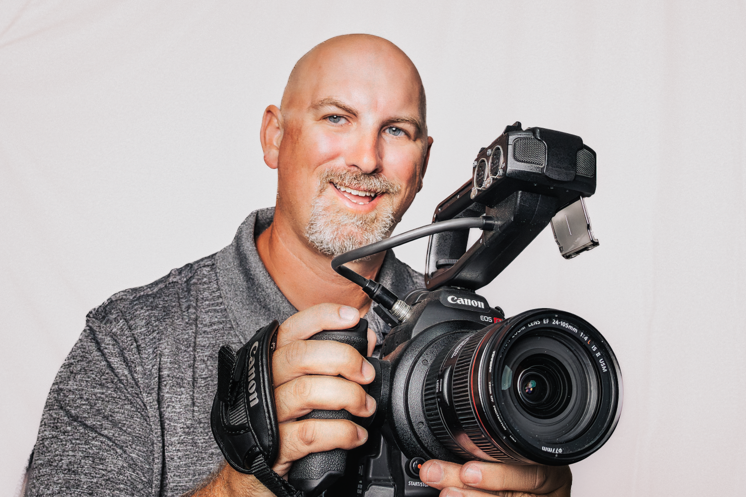 A smiling man holding a professional Canon video camera against a plain white background.