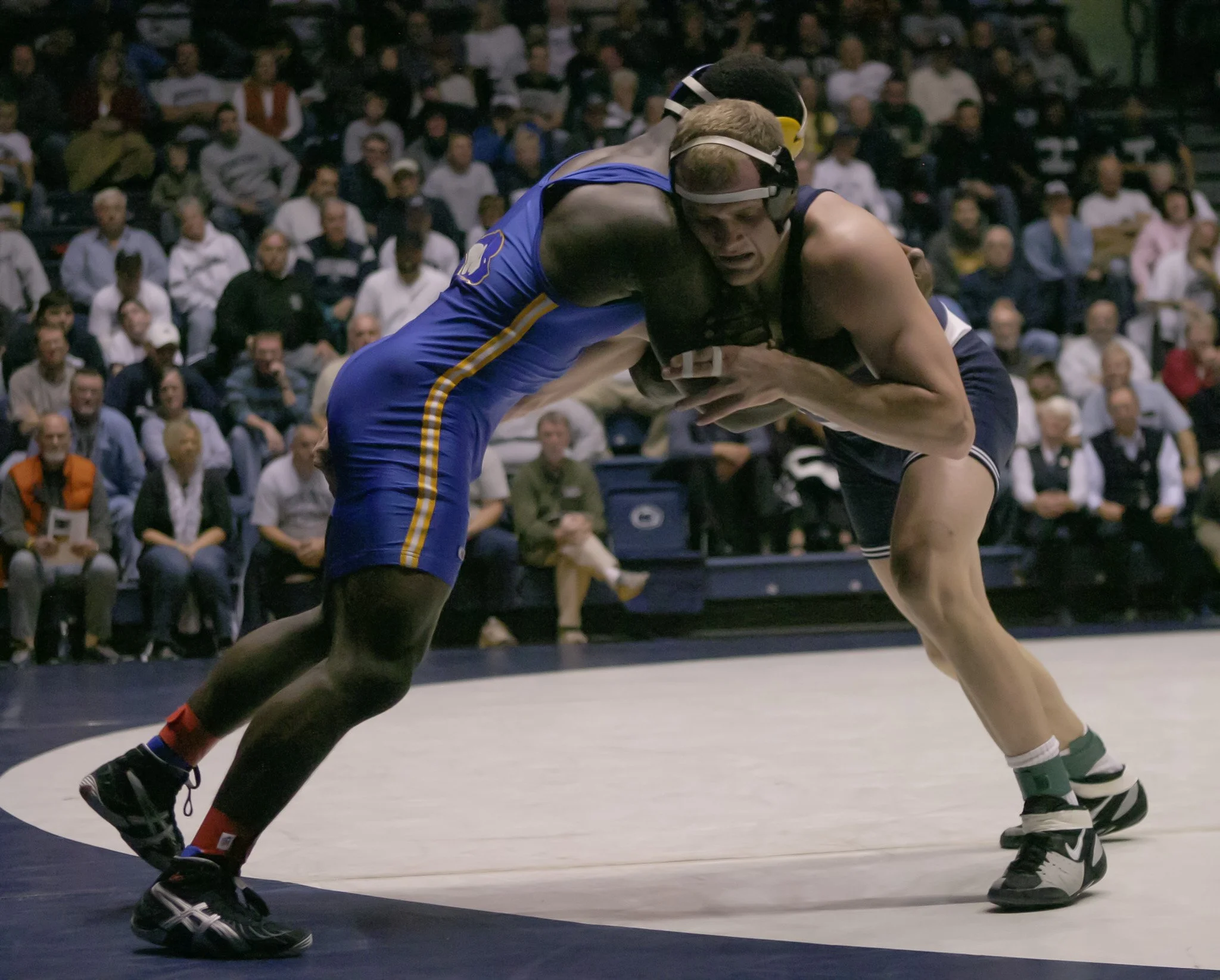 Two wrestlers engaged in a match, one in a blue singlet and the other in a black singlet, with an audience watching them.