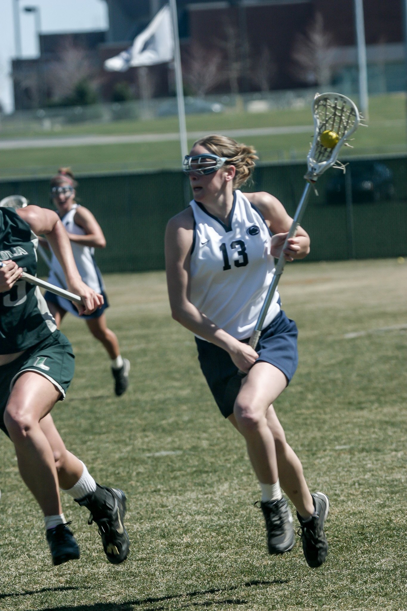 Women playing lacrosse on a grassy field, wearing protective eyewear, with one holding a lacrosse stick with a yellow ball.