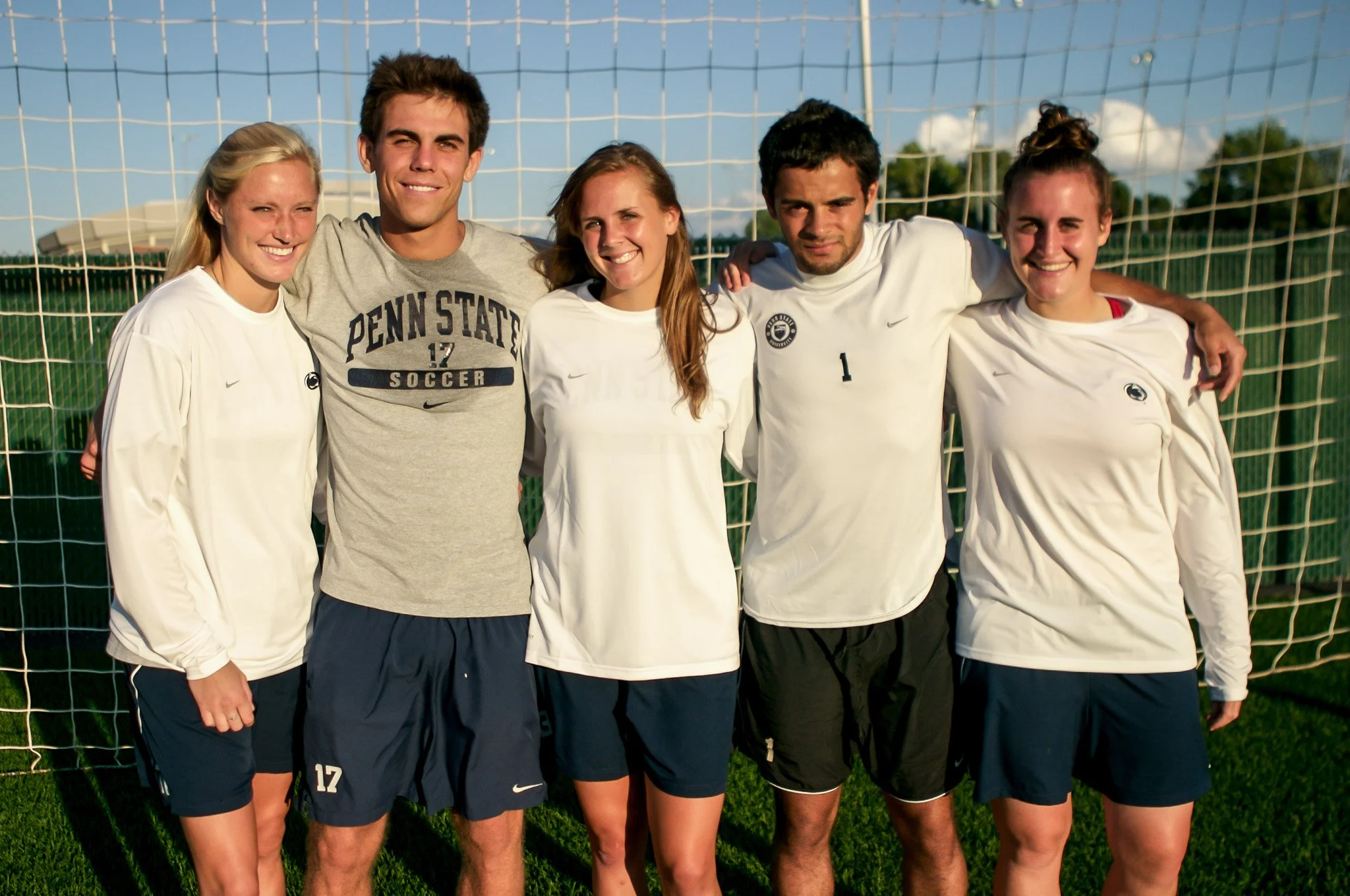 Group of five young adults standing in front of a soccer goal on a grassy field, smiling, two women and three men, dressed in sportswear.