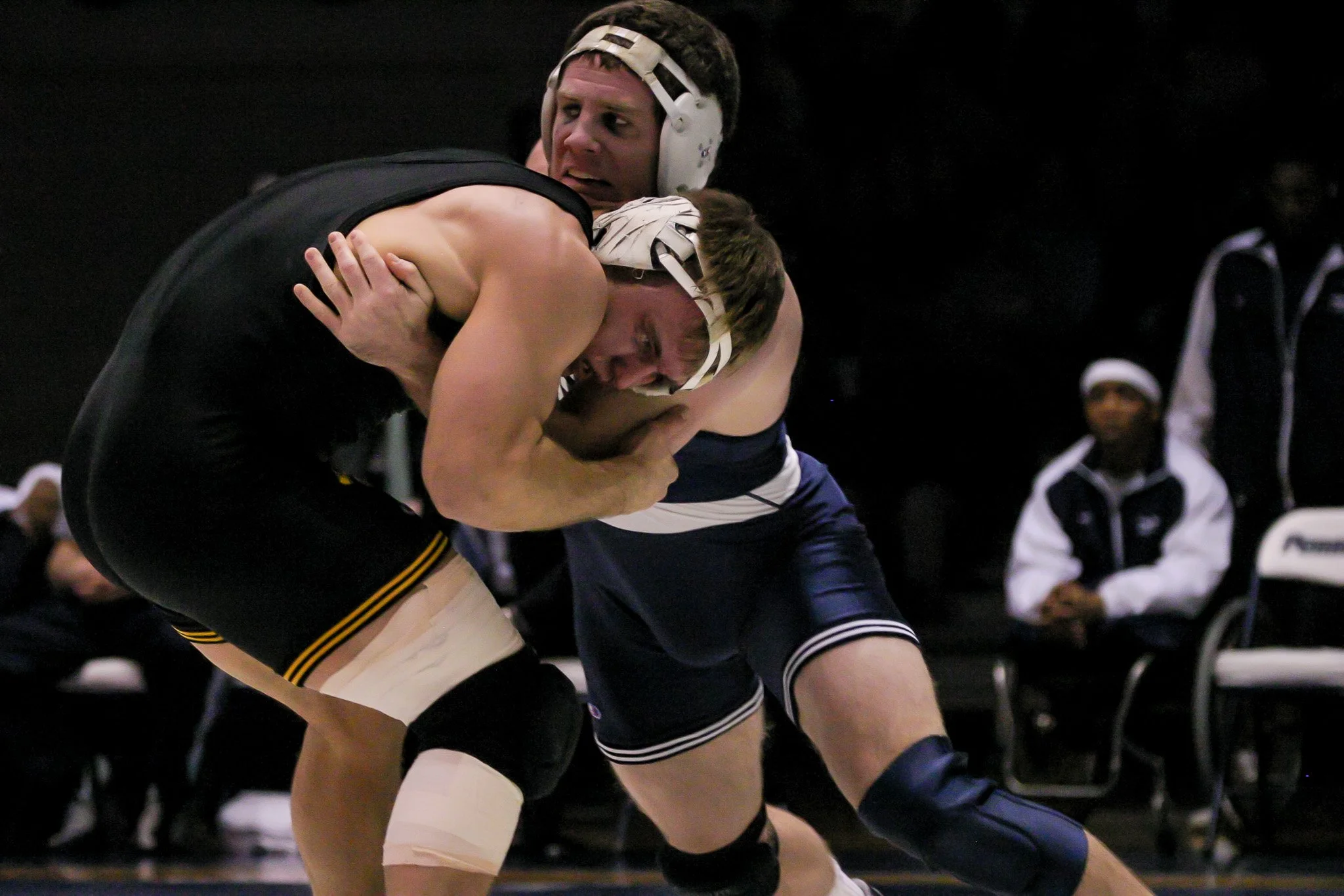 Two wrestlers in a match, one in a black singlet and the other in a blue singlet, grappling on the mat amid spectators.