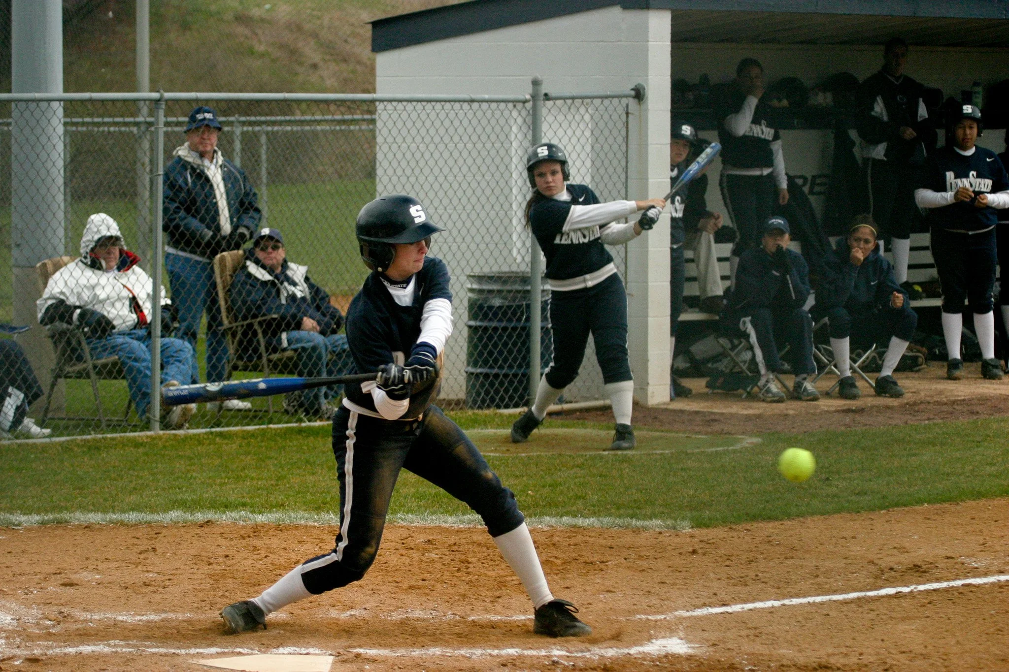 A female softball player swings a bat at a pitch during a game, with teammates and spectators in the background.