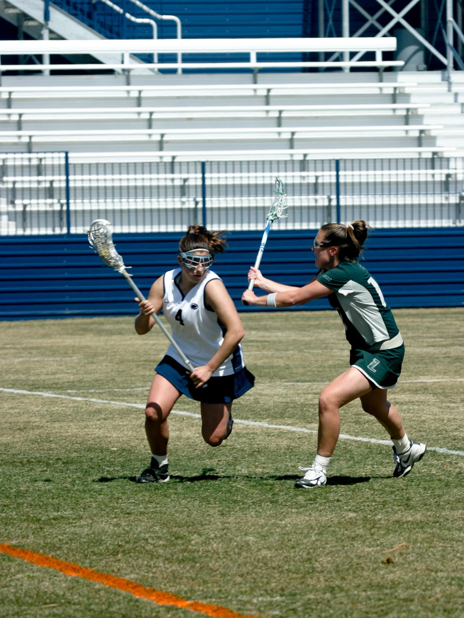 Two female lacrosse players in action on a grassy field, one in a white jersey and the other in a green jersey, competing for the ball.