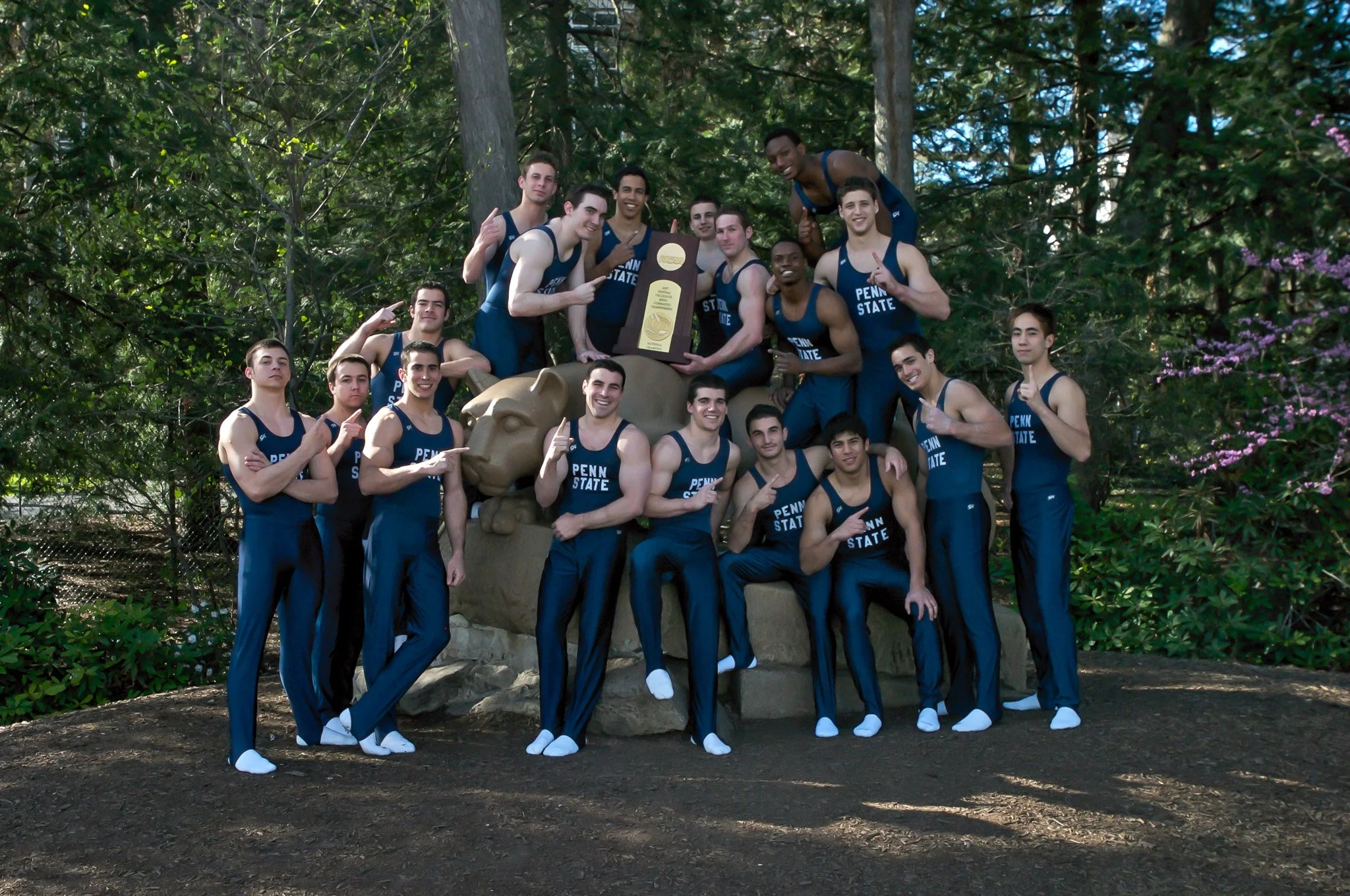 Penn State men's track team posing outdoors with a trophy on a large rock sculpture of a lion's head, surrounded by trees and greenery.