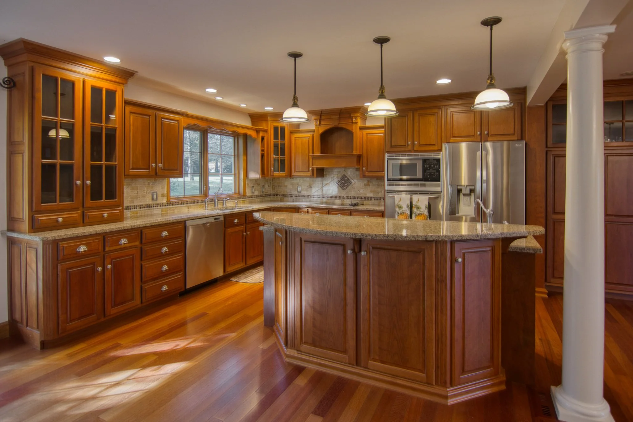 A spacious kitchen with wooden cabinets, a central island, granite countertops, and stainless steel appliances, including a refrigerator and microwave. There is a window above the sink and pendant lights hanging from the ceiling.