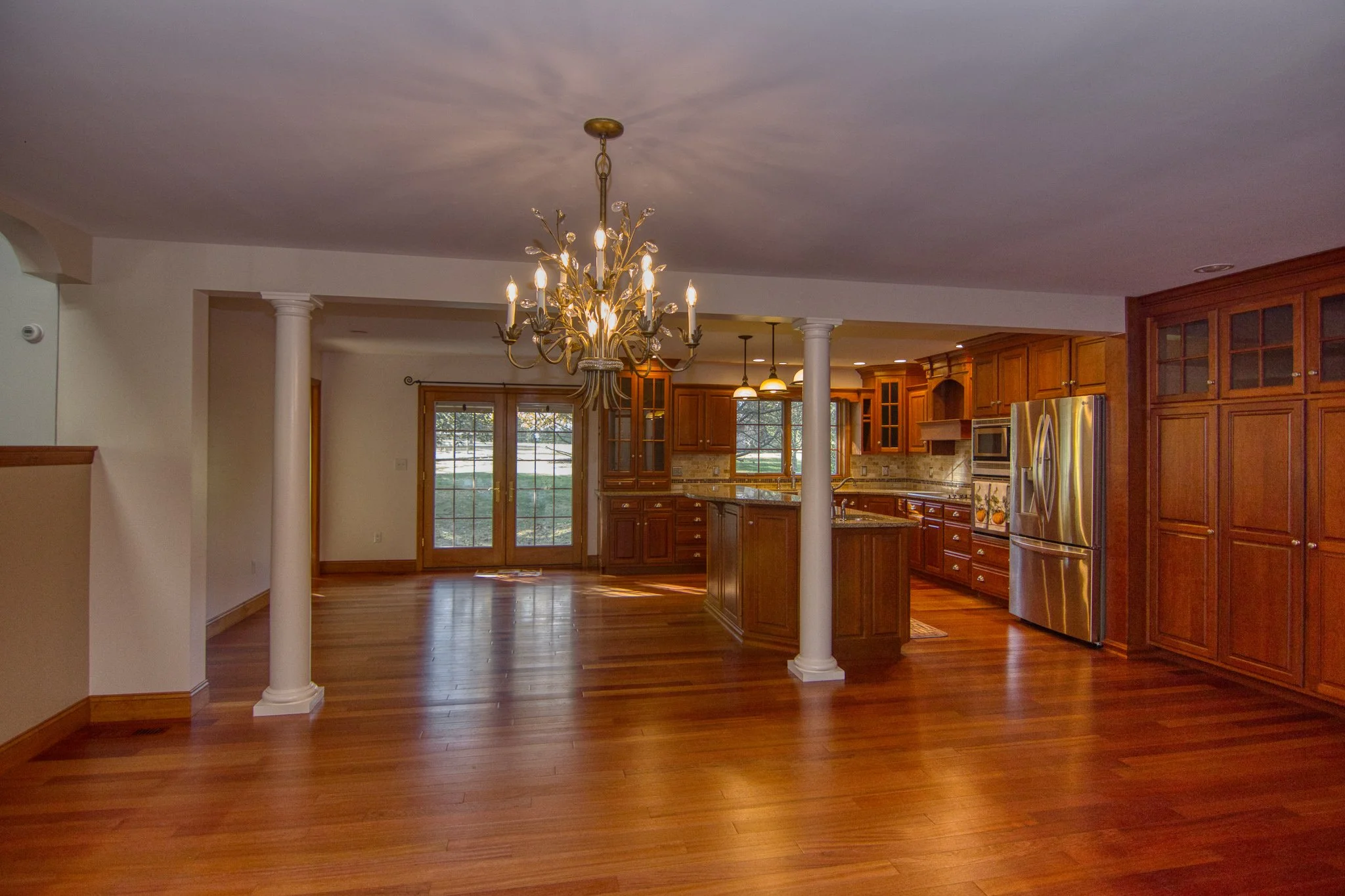 Open-concept kitchen with wooden cabinets, stainless steel appliances, granite countertops, hardwood flooring, a chandelier, and a view of patio doors leading outside.