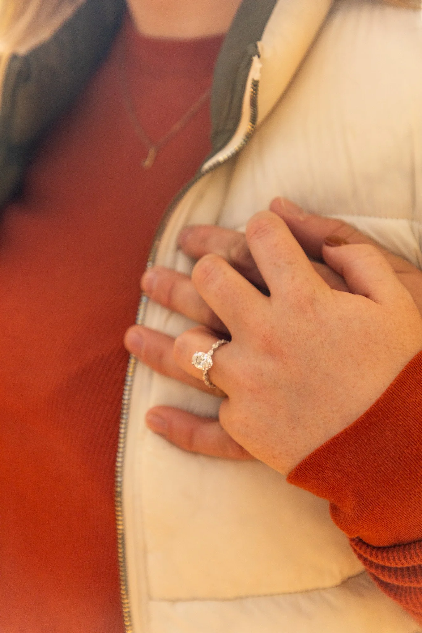 Close-up of a person's hand with a diamond engagement ring, resting on their chest against a beige vest and orange long-sleeve shirt.