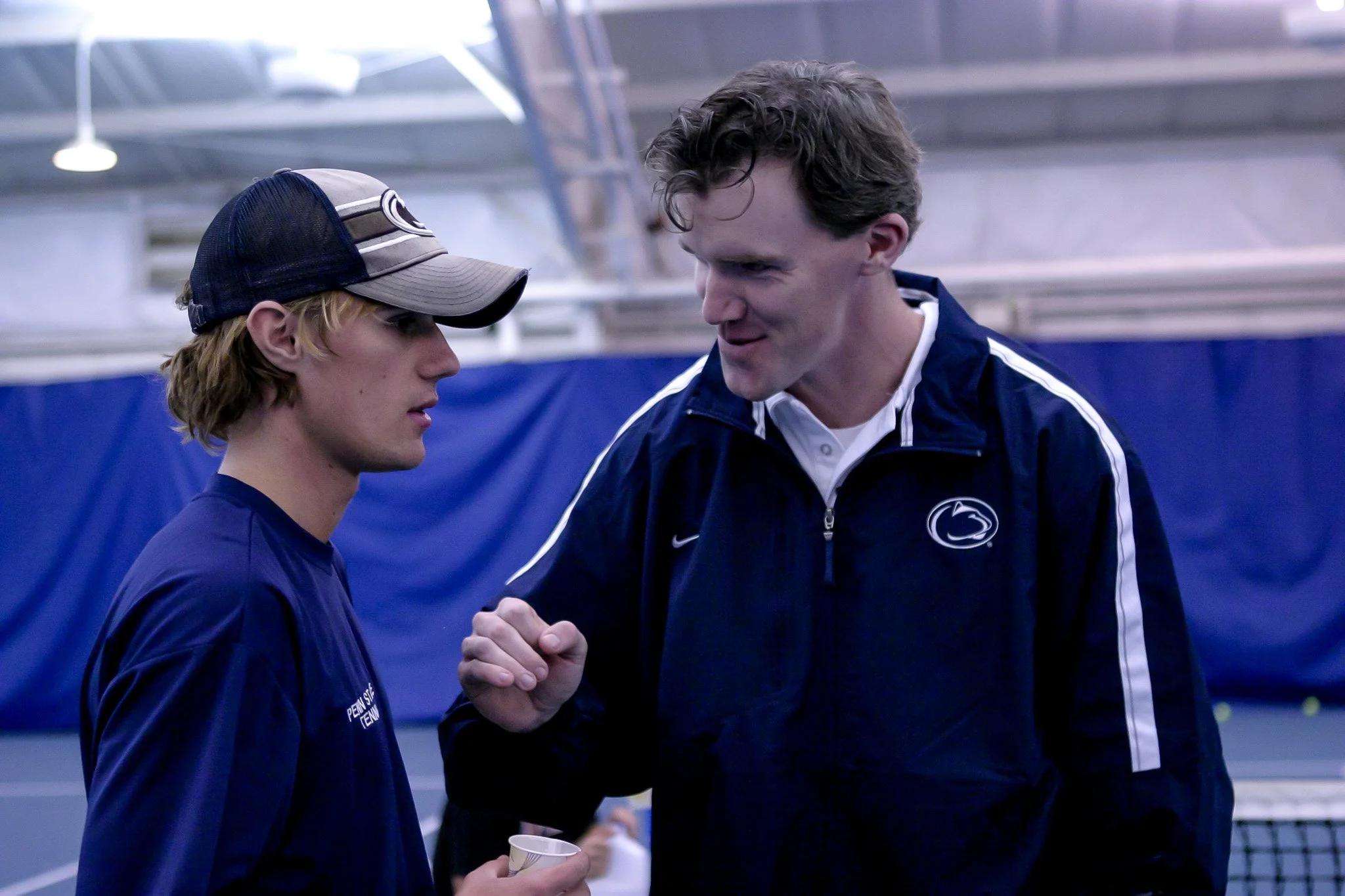 Two men, one younger with blonde hair wearing a cap and Penn State tennis shirt, and an older man with dark hair wearing a Penn State jacket, engaging in a serious conversation on an indoor tennis court.