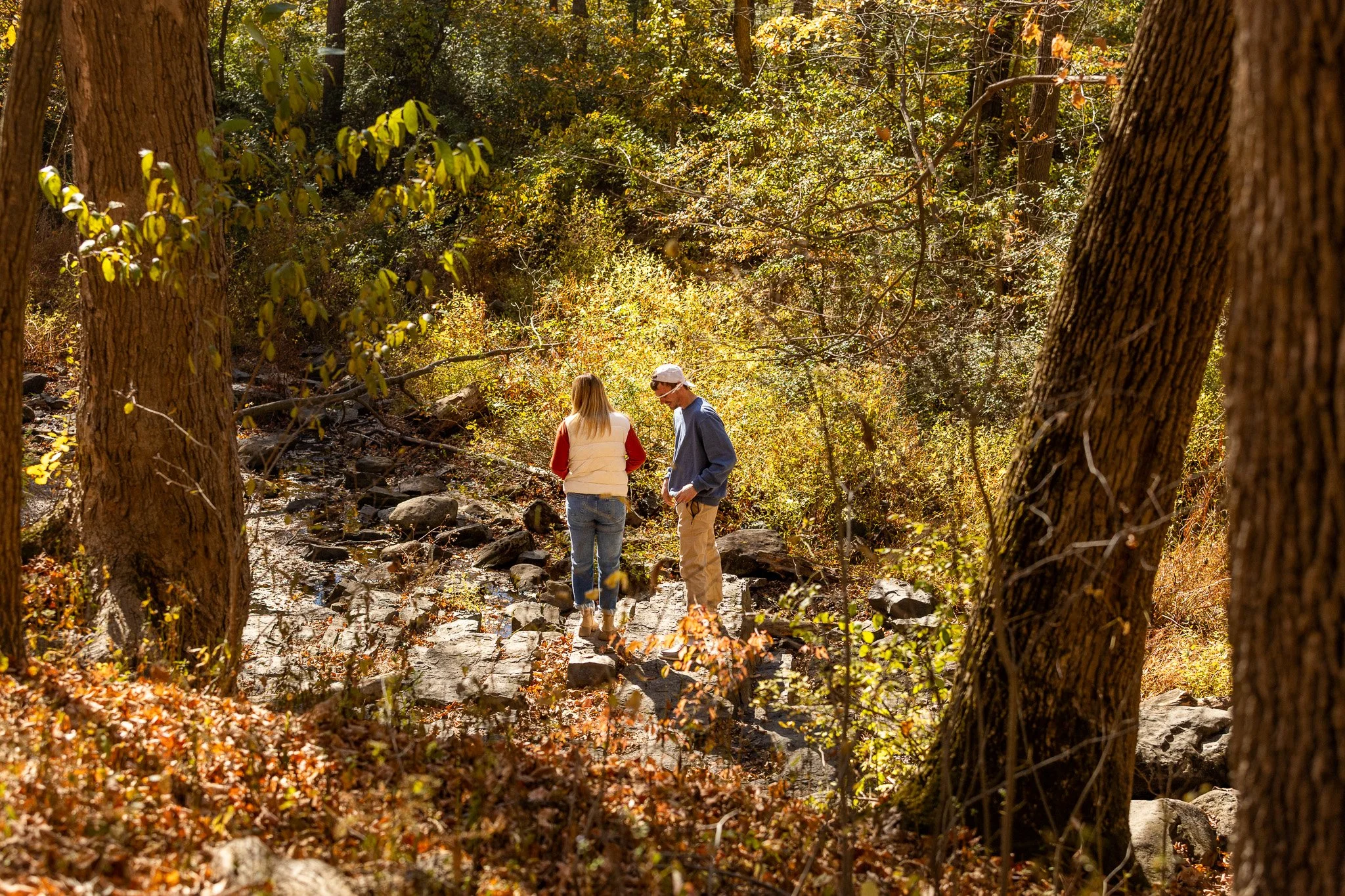 Two people walk through a wooded area with fallen leaves and rocks.