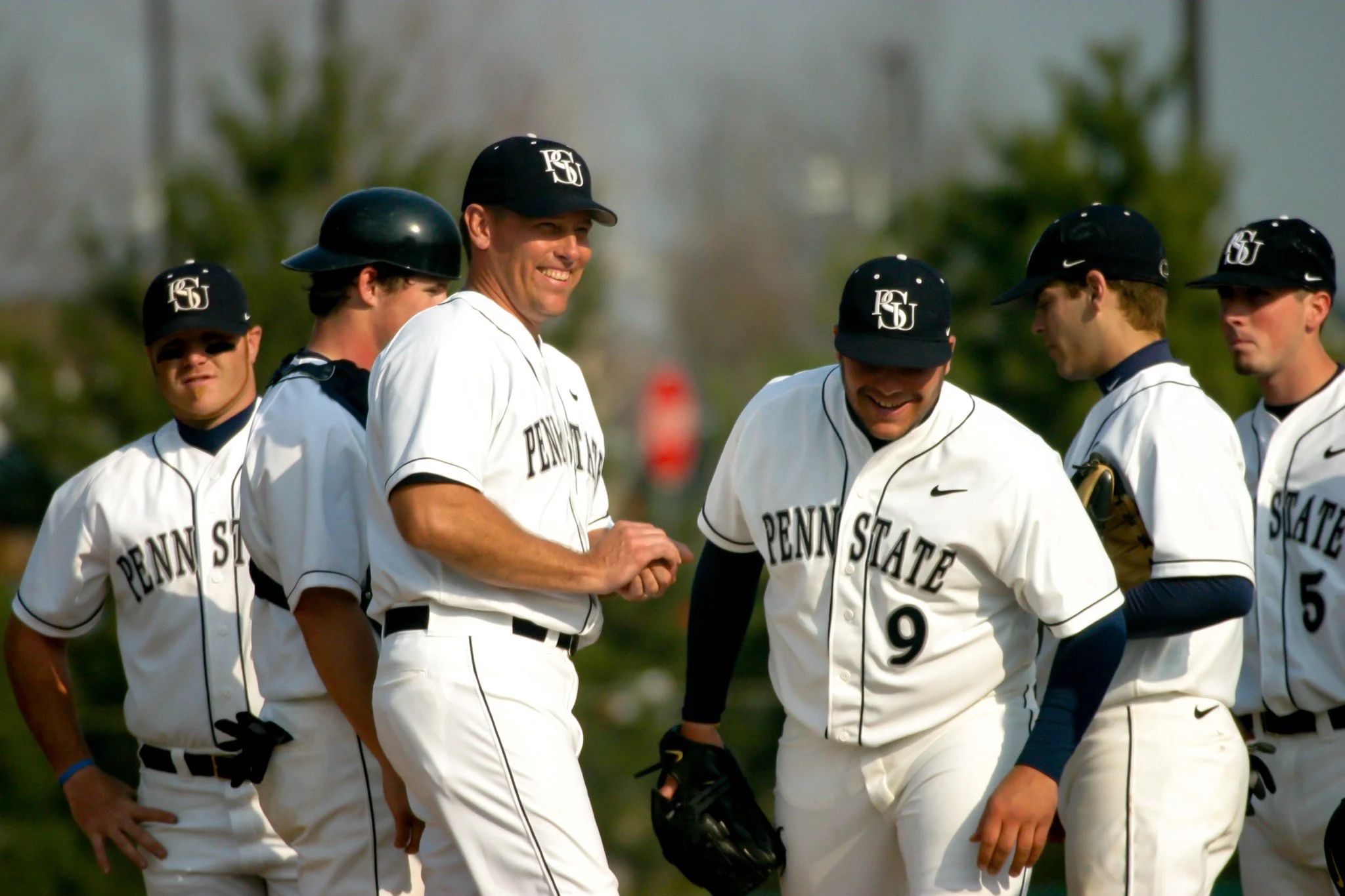 Penn State baseball players in white uniforms with black caps, standing and smiling outdoors on a sunny day.