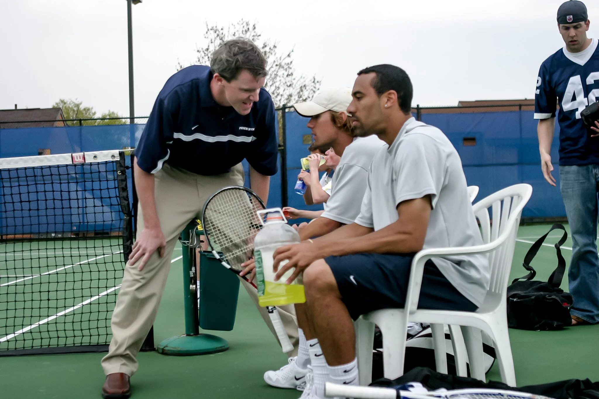 A tennis coach talking to two seated tennis players on a court. The coach is leaning over with a tennis racket, and the players are sitting on white plastic chairs, one holding a water bottle. There is a tennis net and a blue fence behind them.