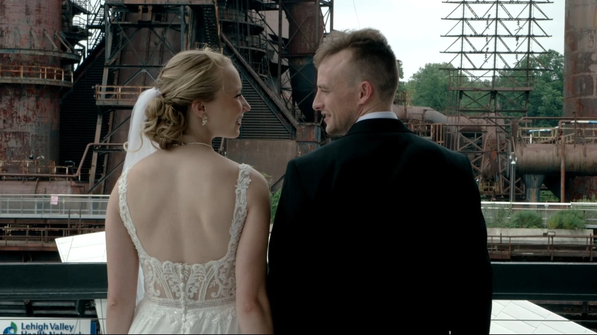 A bride and groom face each other and smile on a balcony with an industrial background of rusted metal structures and pipes during their wedding.