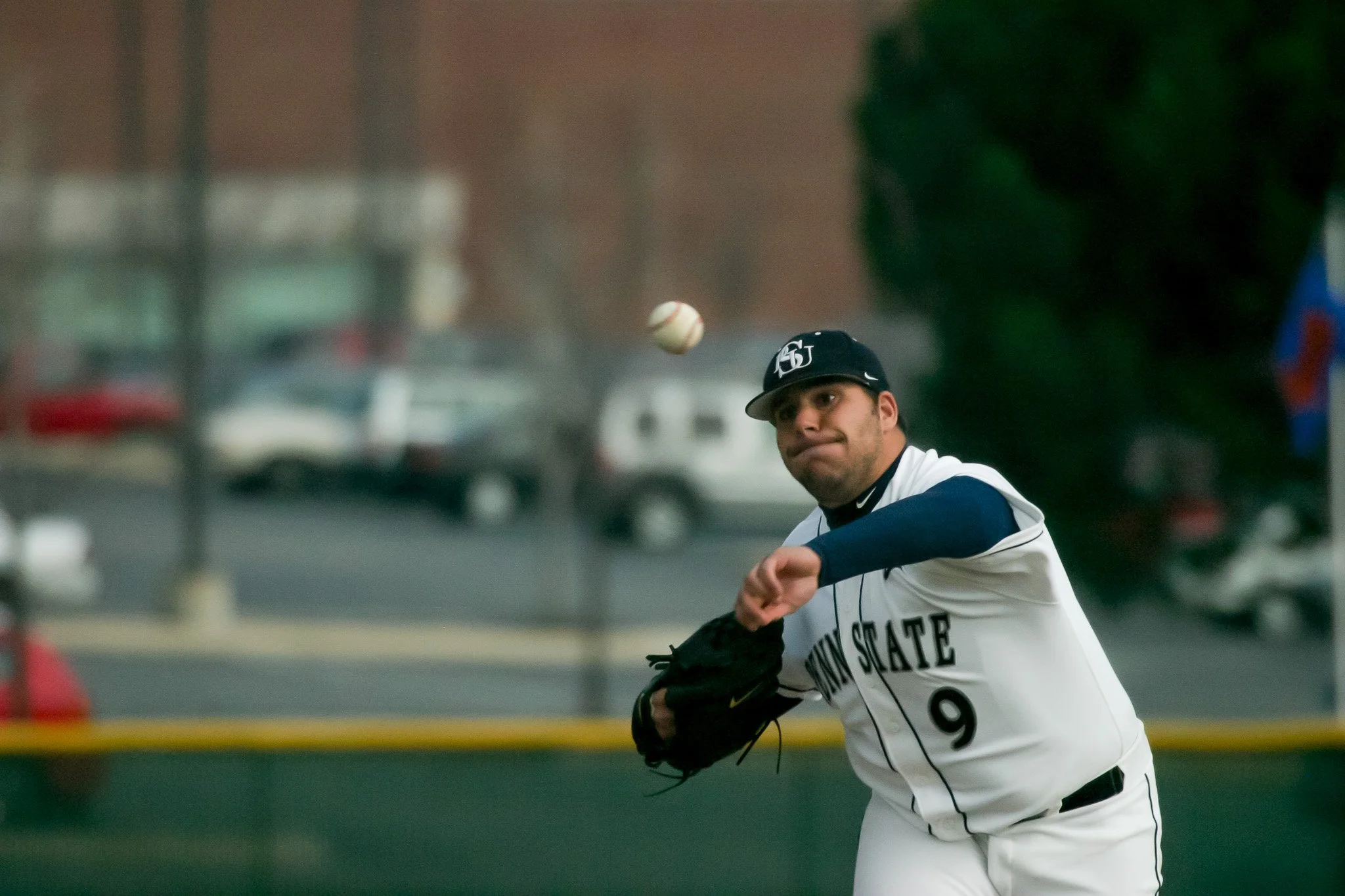 A baseball player from Penn State in white uniform with black and blue details, wearing a black cap, is preparing to catch a baseball in mid-air.