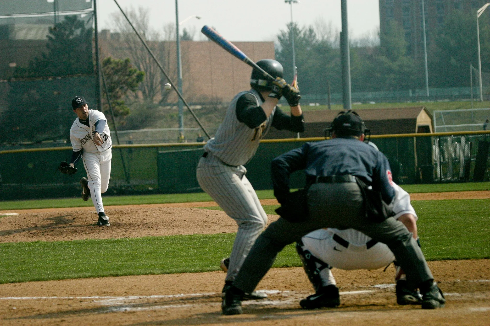 A baseball game in progress with a pitcher in white and navy uniform throwing the ball, a batter wearing a gray pinstripe uniform preparing to swing, an umpire crouching behind the batter, and a catcher in a black and white uniform ready behind home 