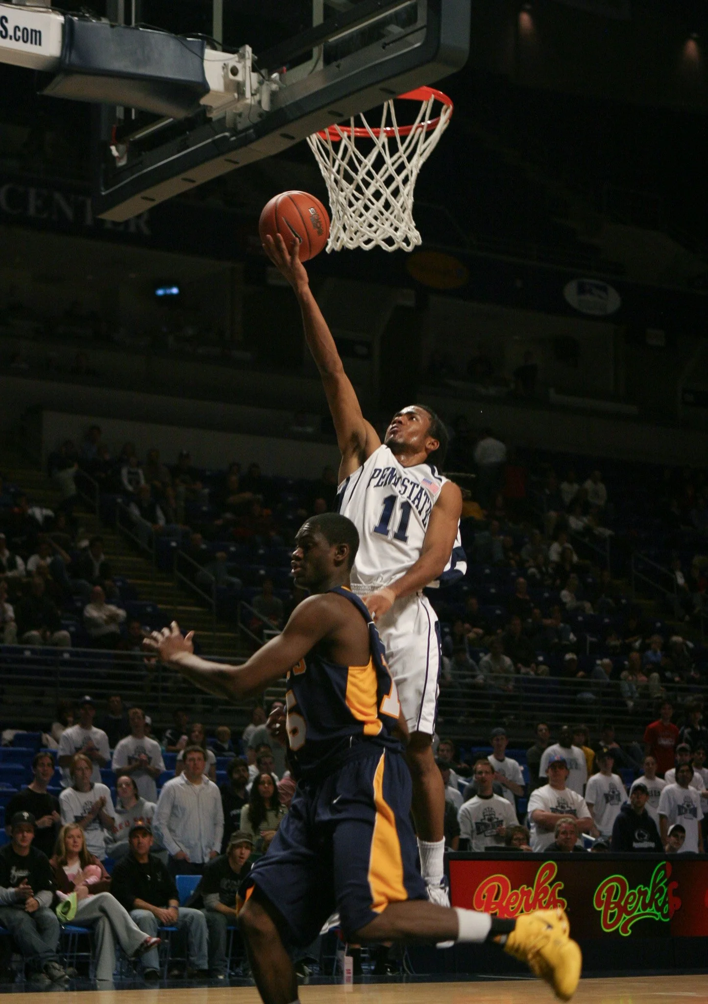 A basketball player in a white jersey with the number 11 is jumping to make a shot, while another player in a dark jersey is attempting to block him. The scene is inside a basketball arena with spectators in the background.