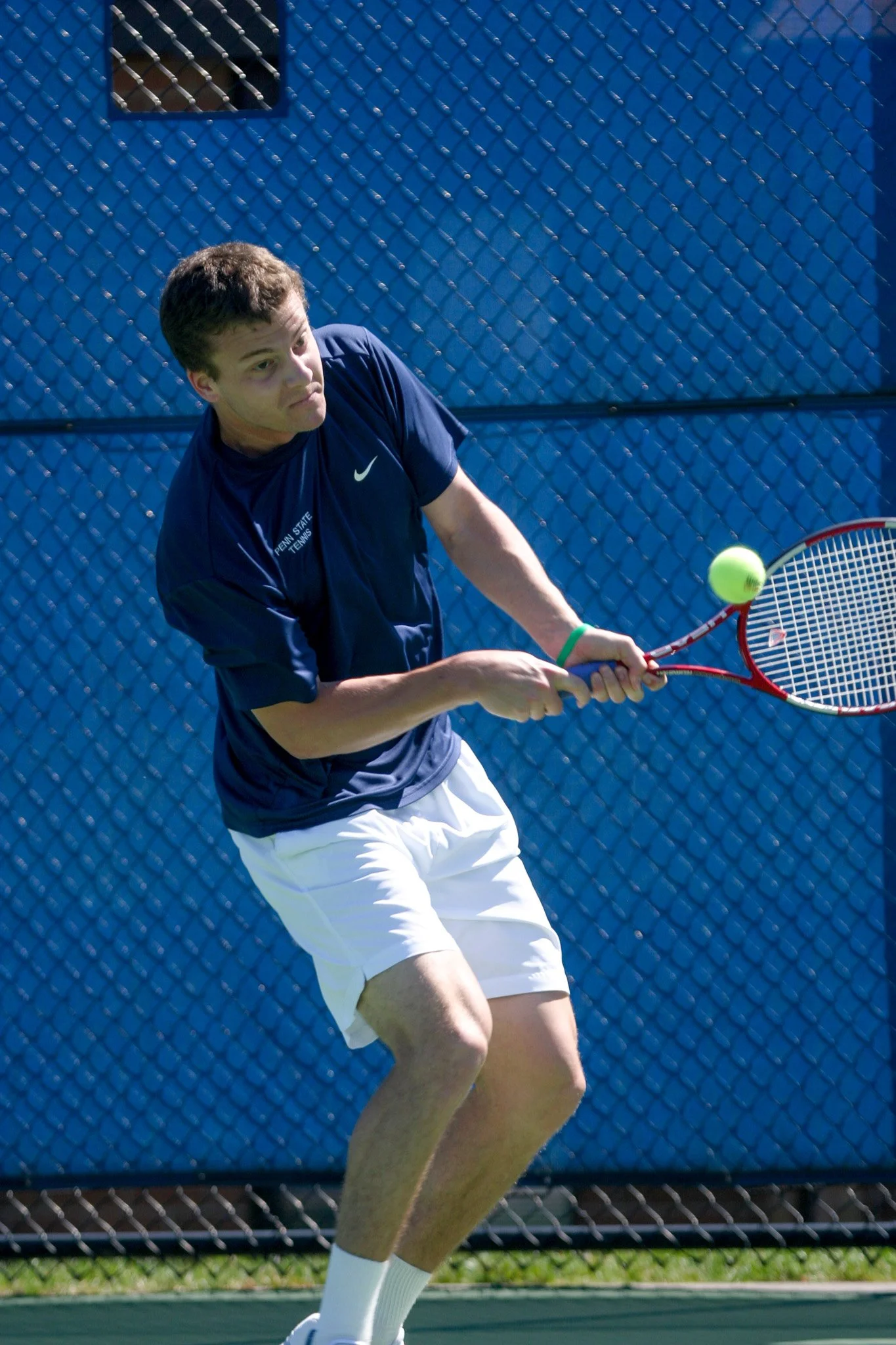 A young man in a navy blue shirt and white shorts playing tennis on a court, preparing to hit a yellow tennis ball with a red tennis racket.