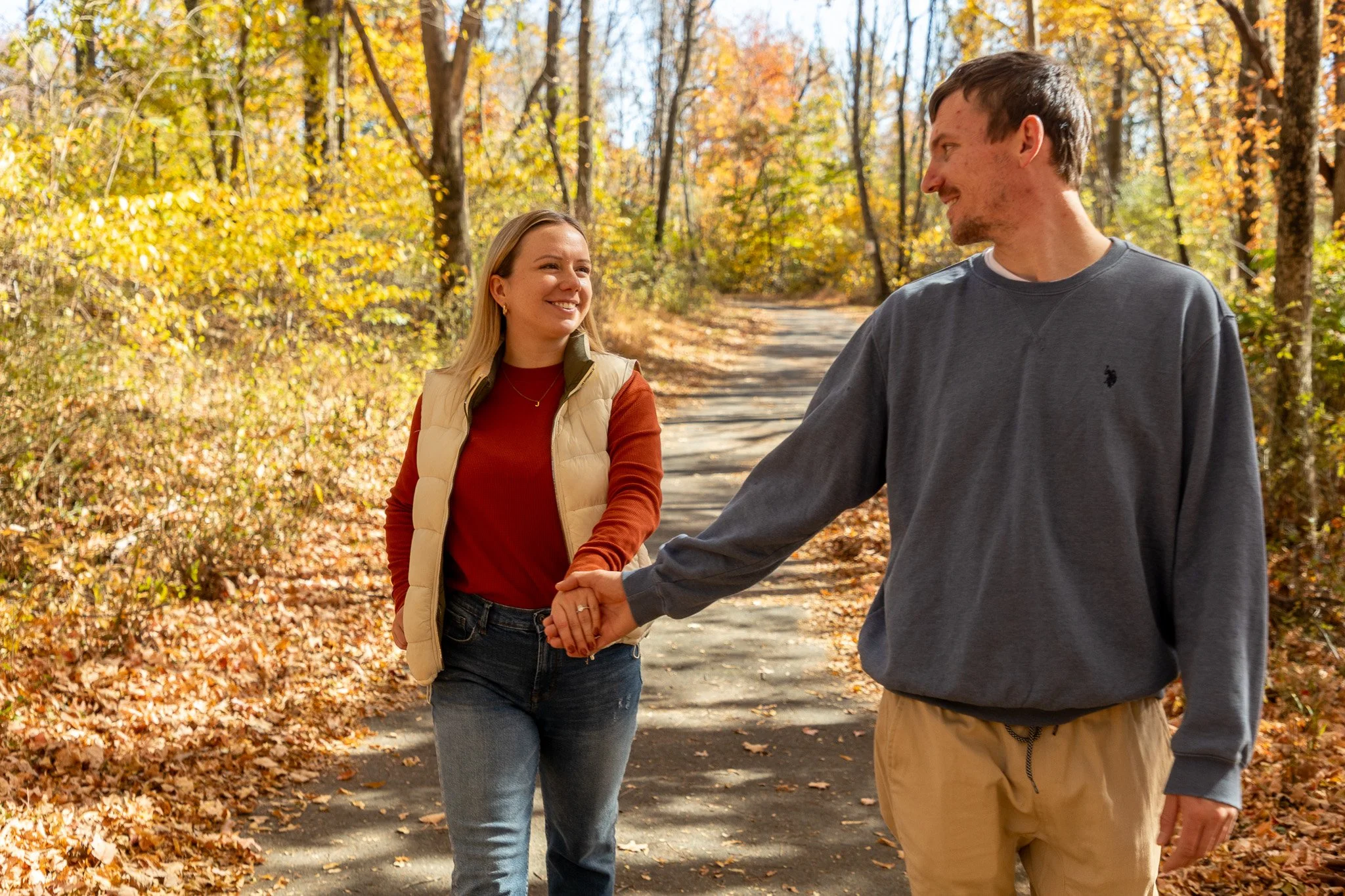 A young woman and a young man holding hands and walking along a forest trail with autumn-colored trees and fallen leaves.
