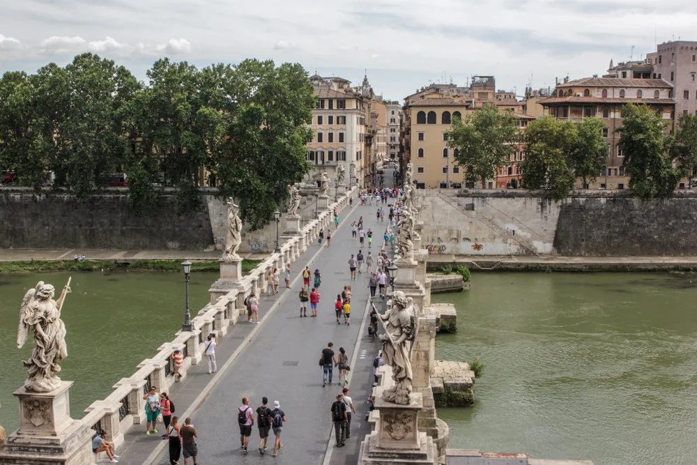 People walking across the Ponte Sant'Angelo bridge in Rome, Italy, lined with statues, with a cityscape and trees in the background.