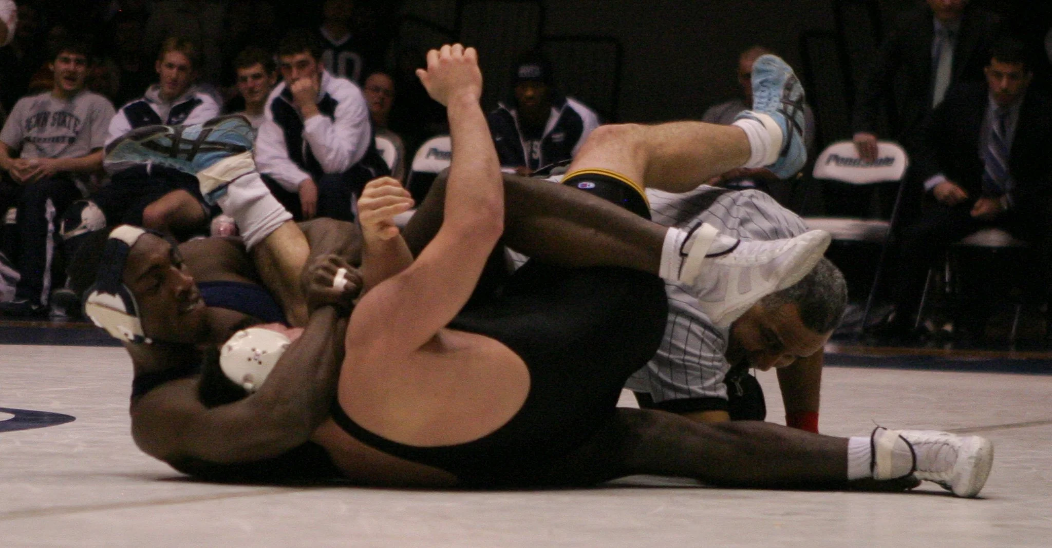 Two wrestlers compete on a wrestling mat while a referee watches. One wrestler, wearing black, is on the ground holding the other wrestler, who is in a light-colored uniform, in a pin.