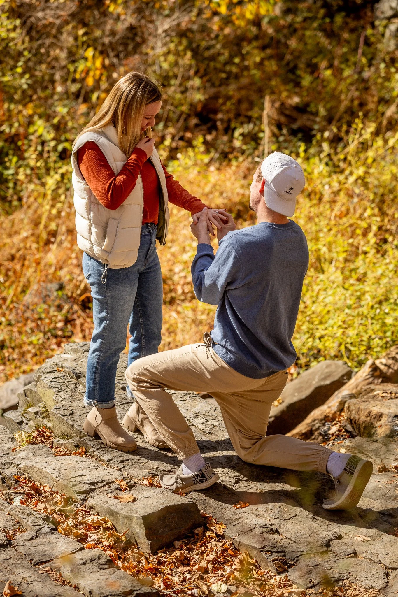 A man proposing marriage to a woman on a rocky trail in autumn, with fallen leaves and trees in the background.