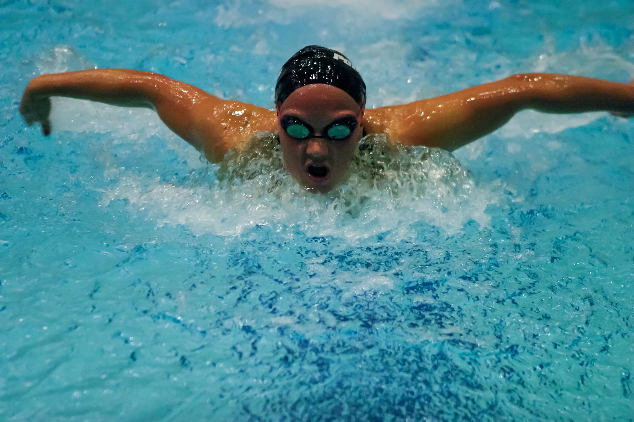 A swimmer performing the butterfly stroke in a pool, wearing goggles and a swim cap, with their arms extended forward and face submerged in the water.