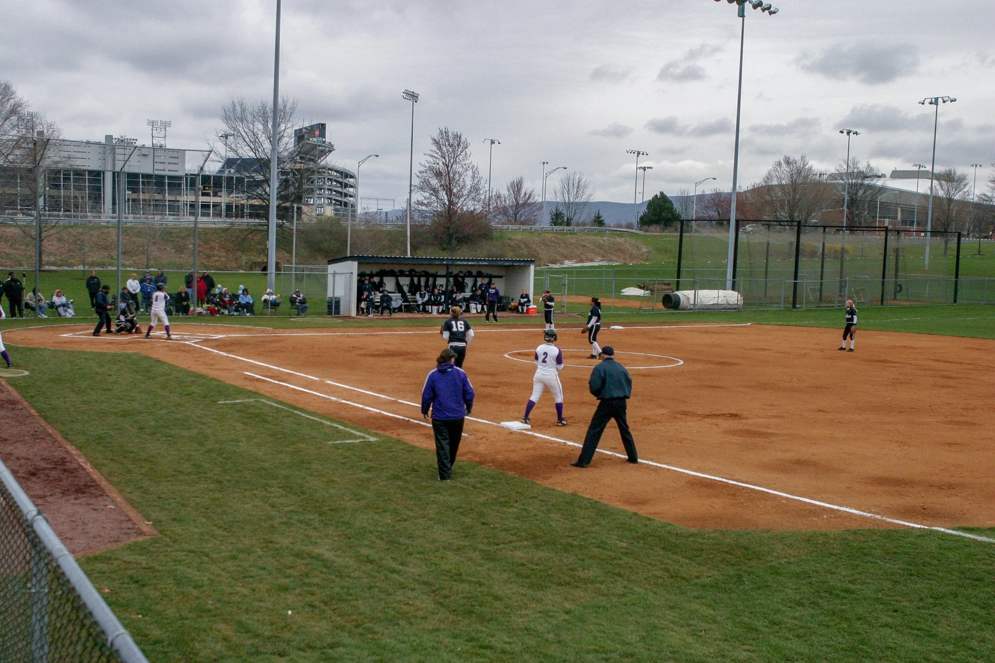 A baseball game is taking place on a cloudy day. Players are on the field and the batter is at home plate. Coaches and spectators are present along the sidelines and sitting on benches.