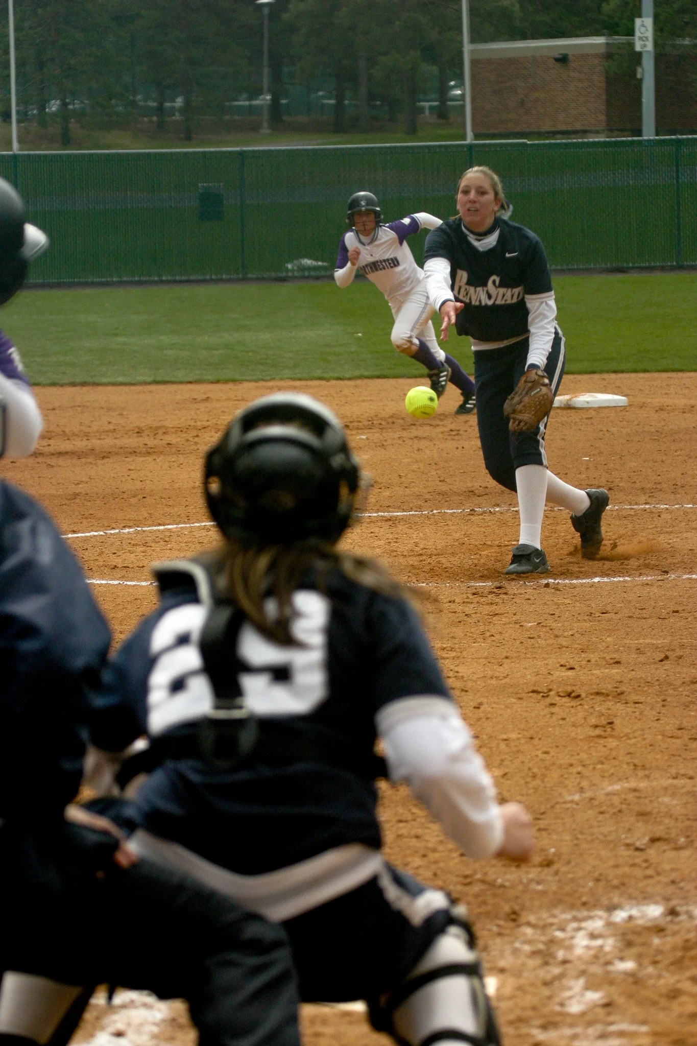 A softball player pitching the ball on a field, with teammates and a catcher in the foreground.