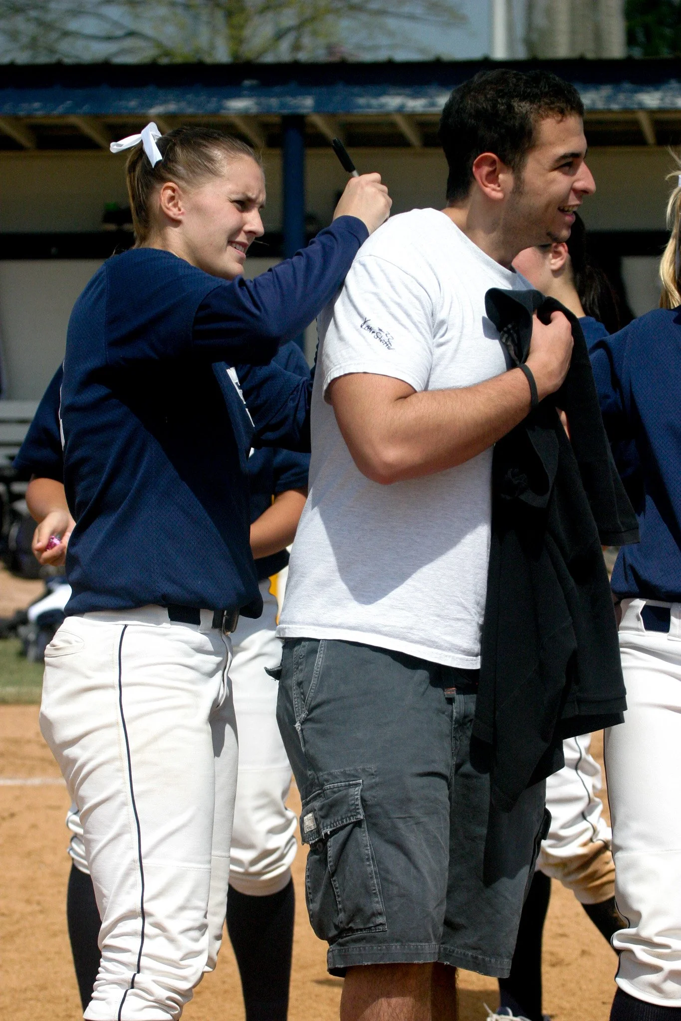 People standing in a line on a baseball field, with one person talking to another. The woman is wearing a navy blue sports jacket and white baseball pants, and the man in front is holding a black jacket and wearing a white t-shirt and dark shorts.