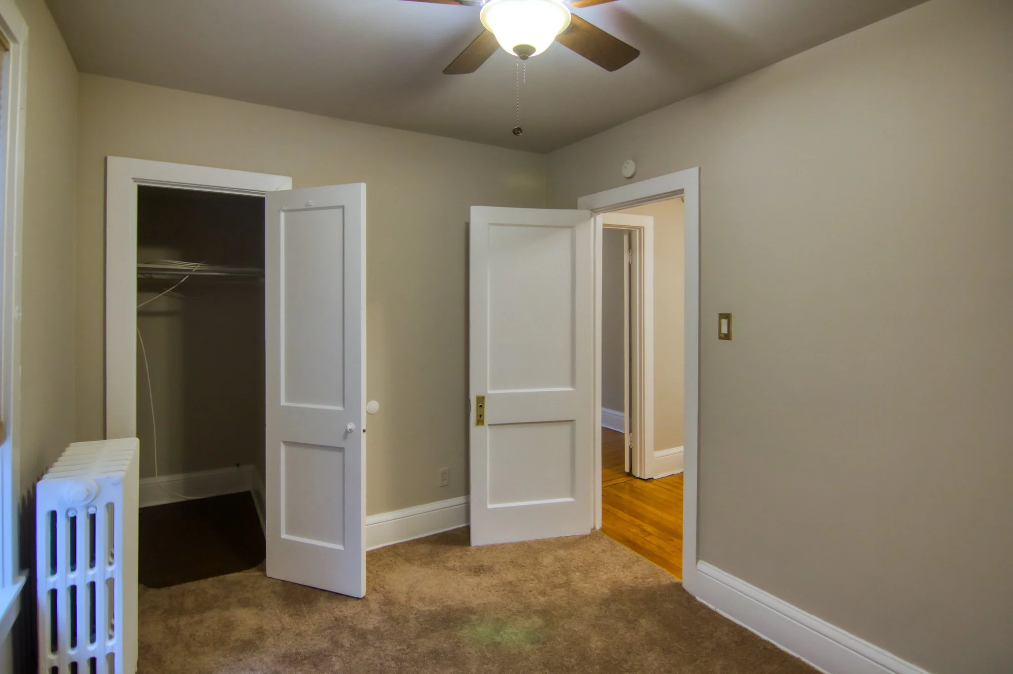 Empty room with closet, open door leading to hallway, ceiling fan, beige walls, carpeted floor, and a radiator.