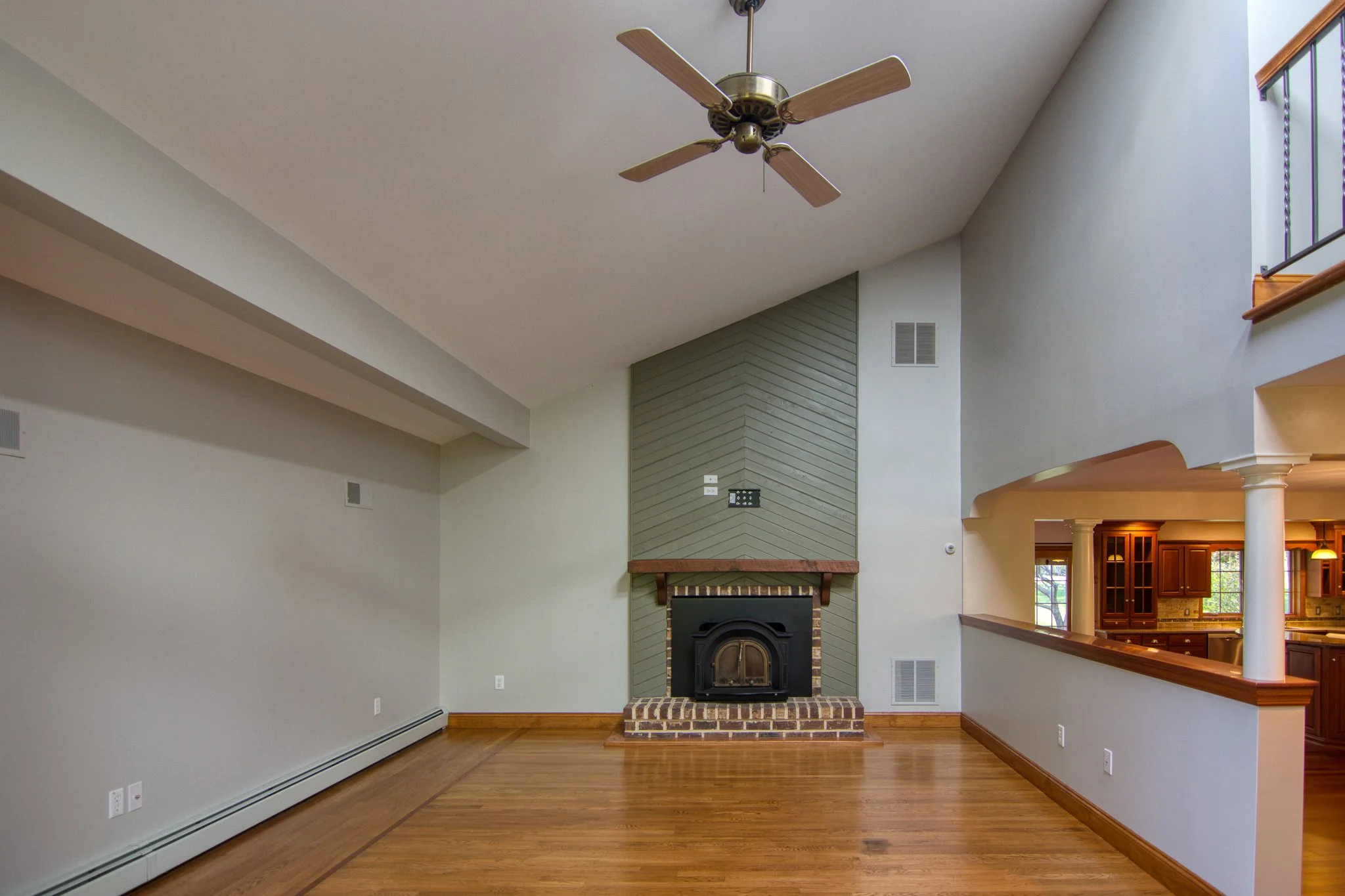 Living room with a brick fireplace, vaulted ceiling with a ceiling fan, hardwood floors, and view into a kitchen area.