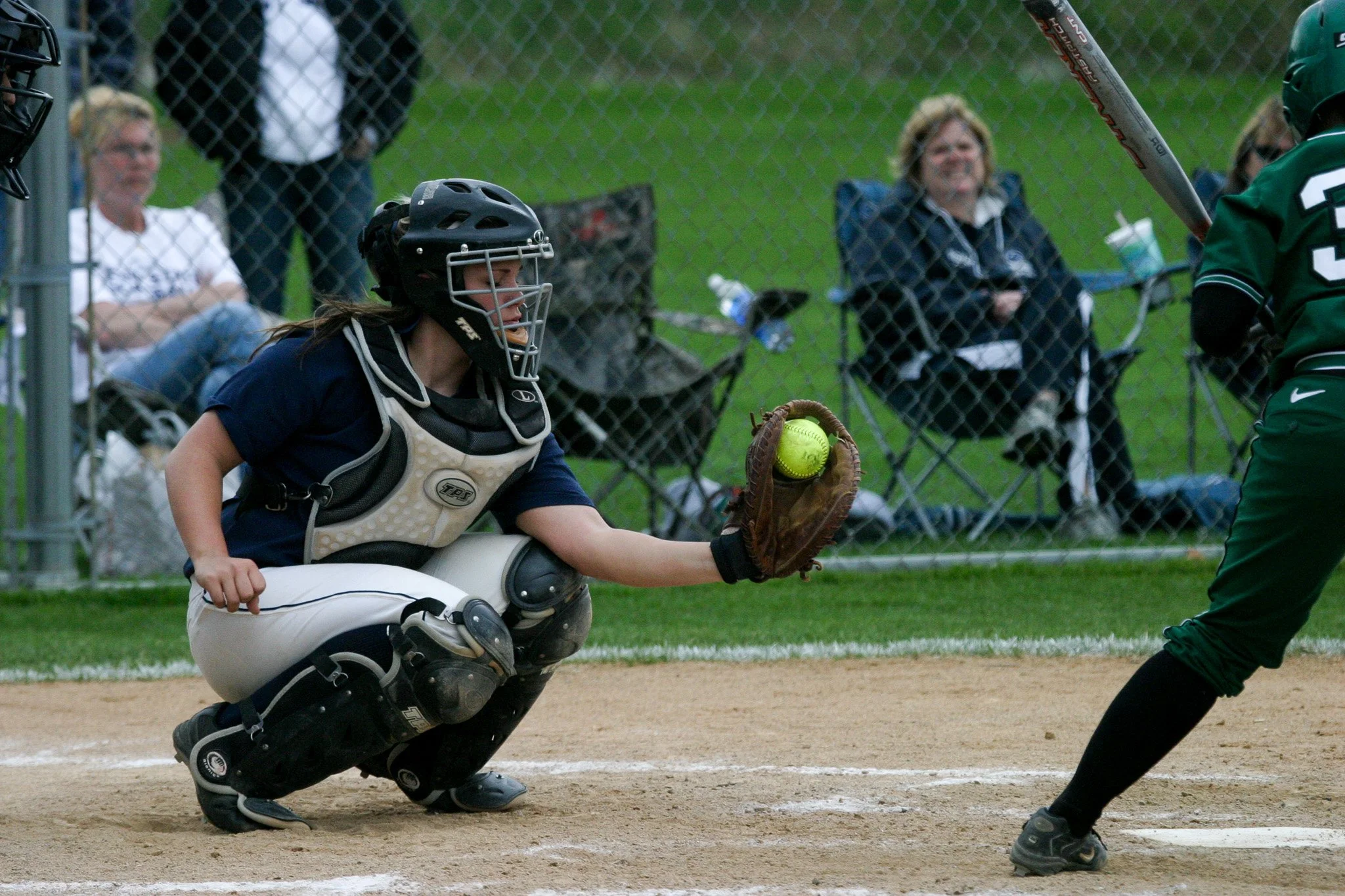 A young girl playing softball, squatting in a catcher's position, wearing a helmet, catcher's gear, and glove, reaching out to catch a yellow softball.