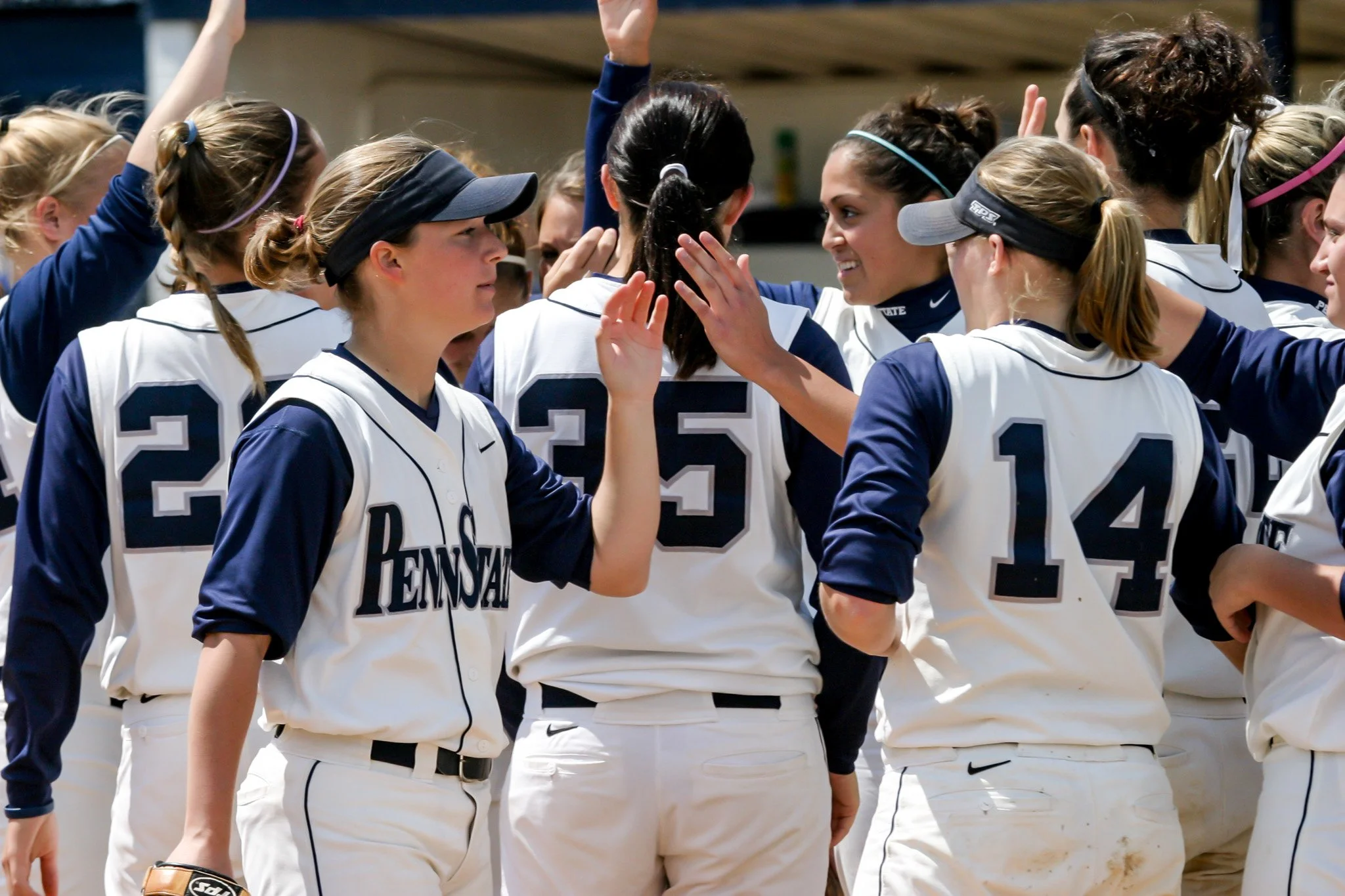 A group of female softball players in white and navy uniforms huddled together, some wearing visors, during a game or practice.