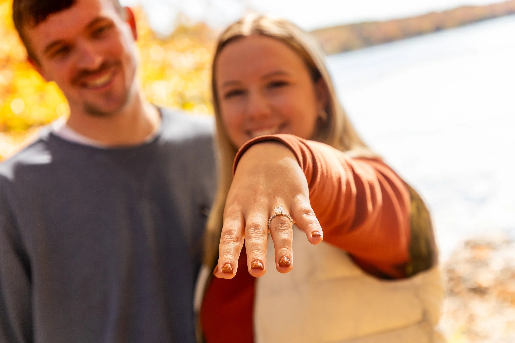 A woman showing her engagement ring on her finger with a smiling man standing behind her outdoors near a body of water with autumn leaves on the trees.