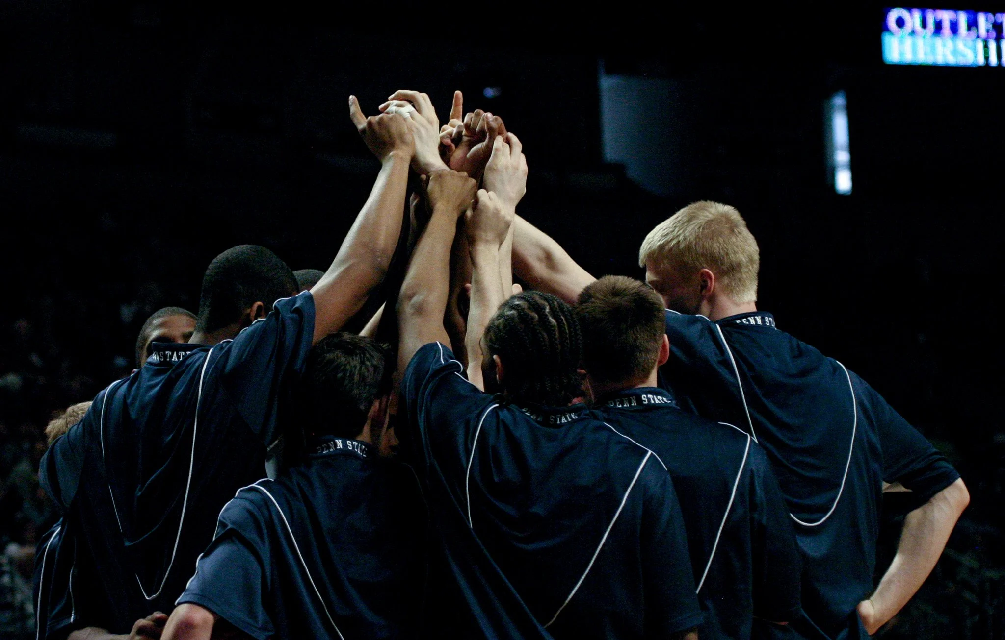 A group of basketball players in blue jerseys huddled together in a team cheer with hands united in the center.