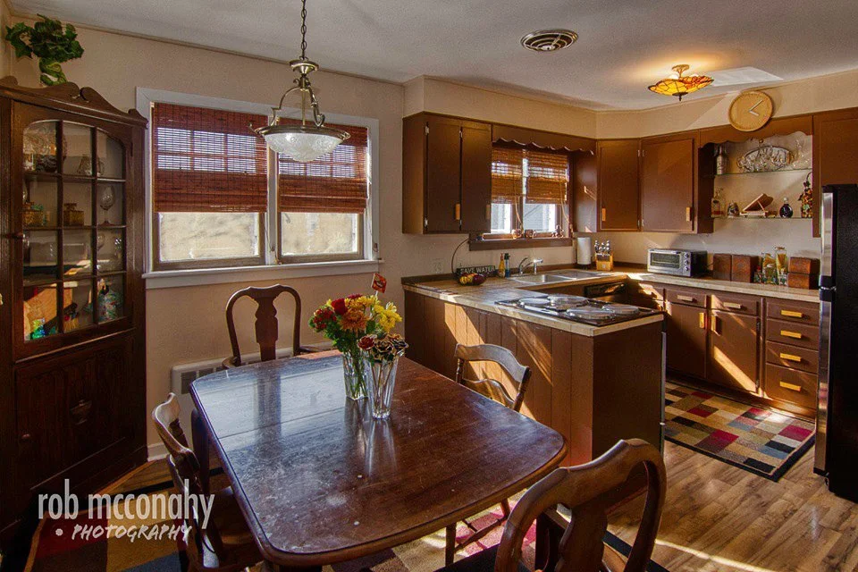 A kitchen and dining area with a wooden table and chairs, a window with blinds, a cabinet, and kitchen appliances, with sunlight streaming in.