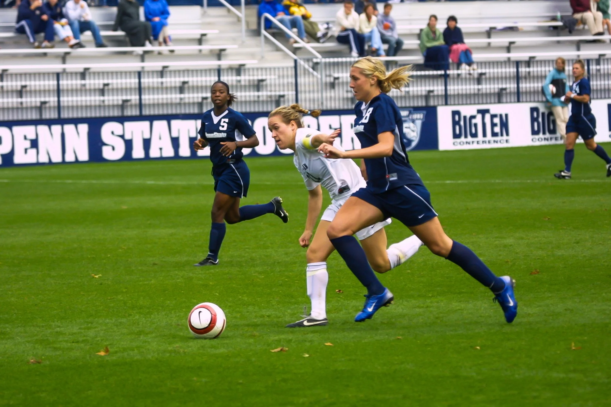 Women’s soccer game on a green field with players in blue and white jerseys, some running after the ball, with spectators in the stands and advertising banners in the background.