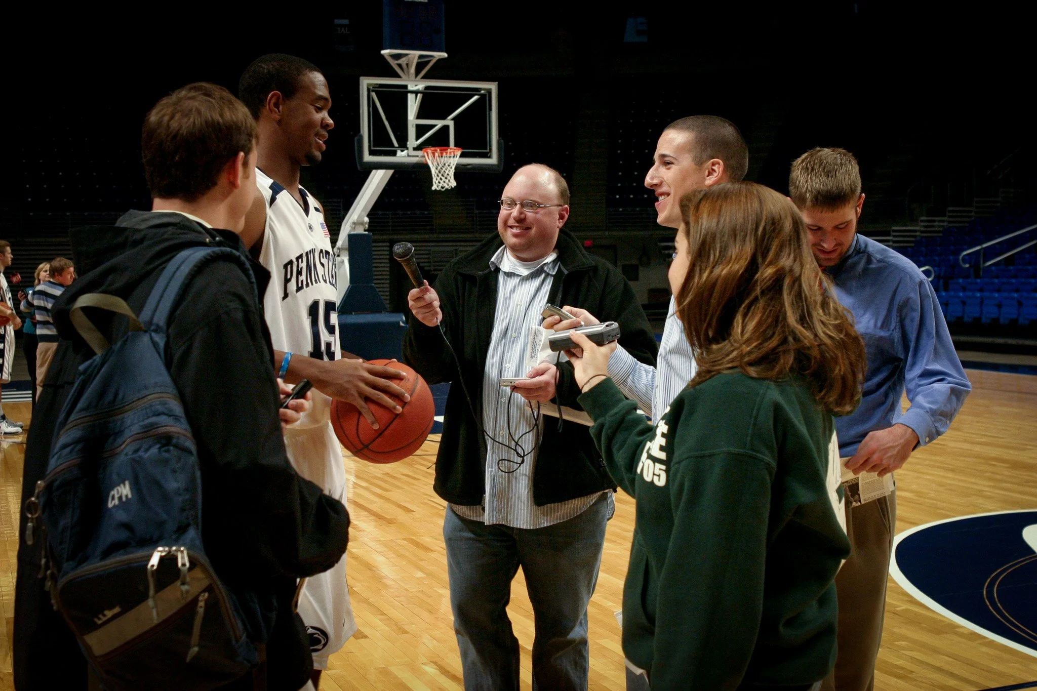 A basketball player in a white Penn State uniform holding a basketball, surrounded by reporters on a basketball court.