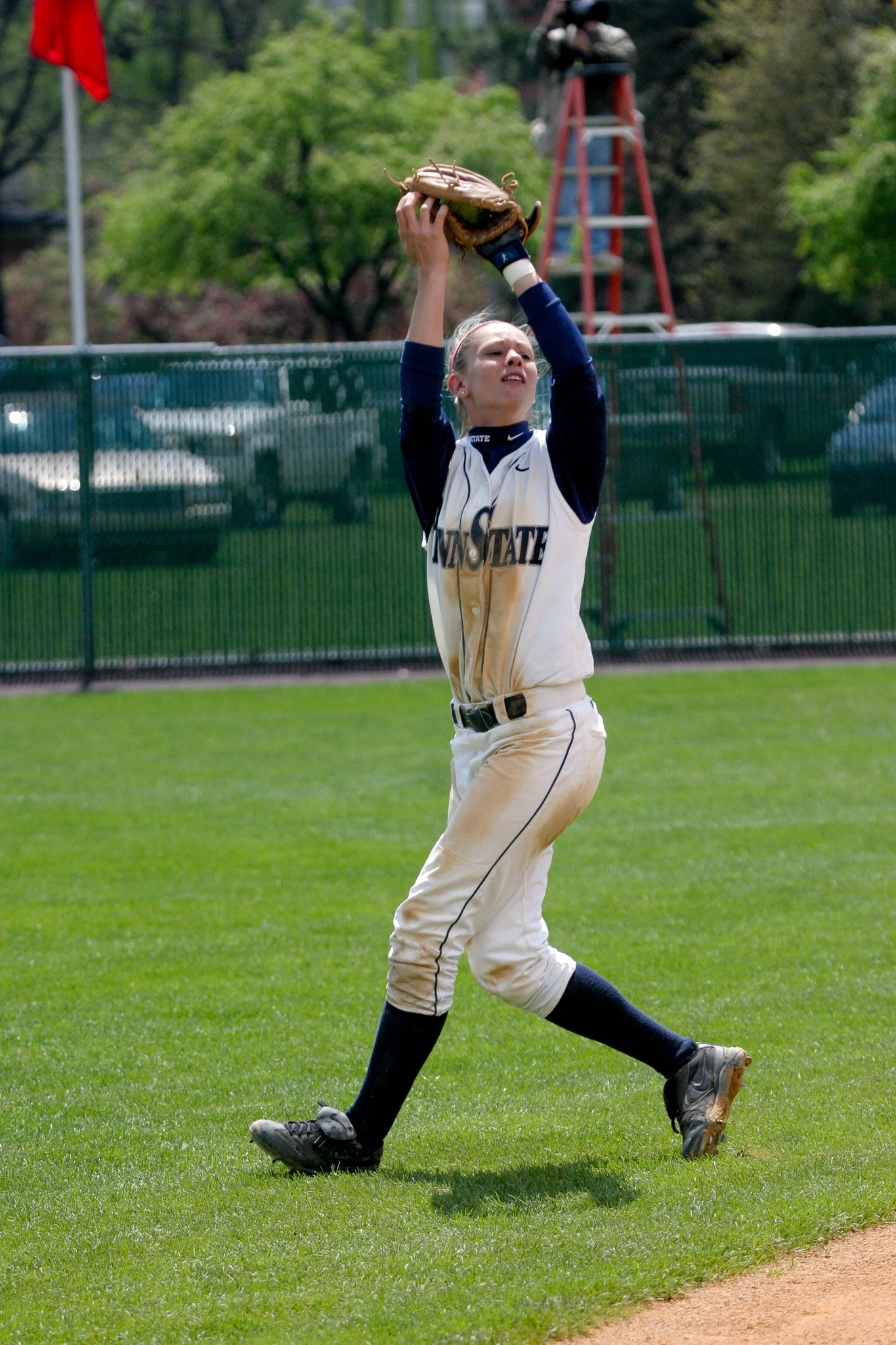 A female softball player in a white and navy uniform catching a fly ball with her glove on a grassy field.