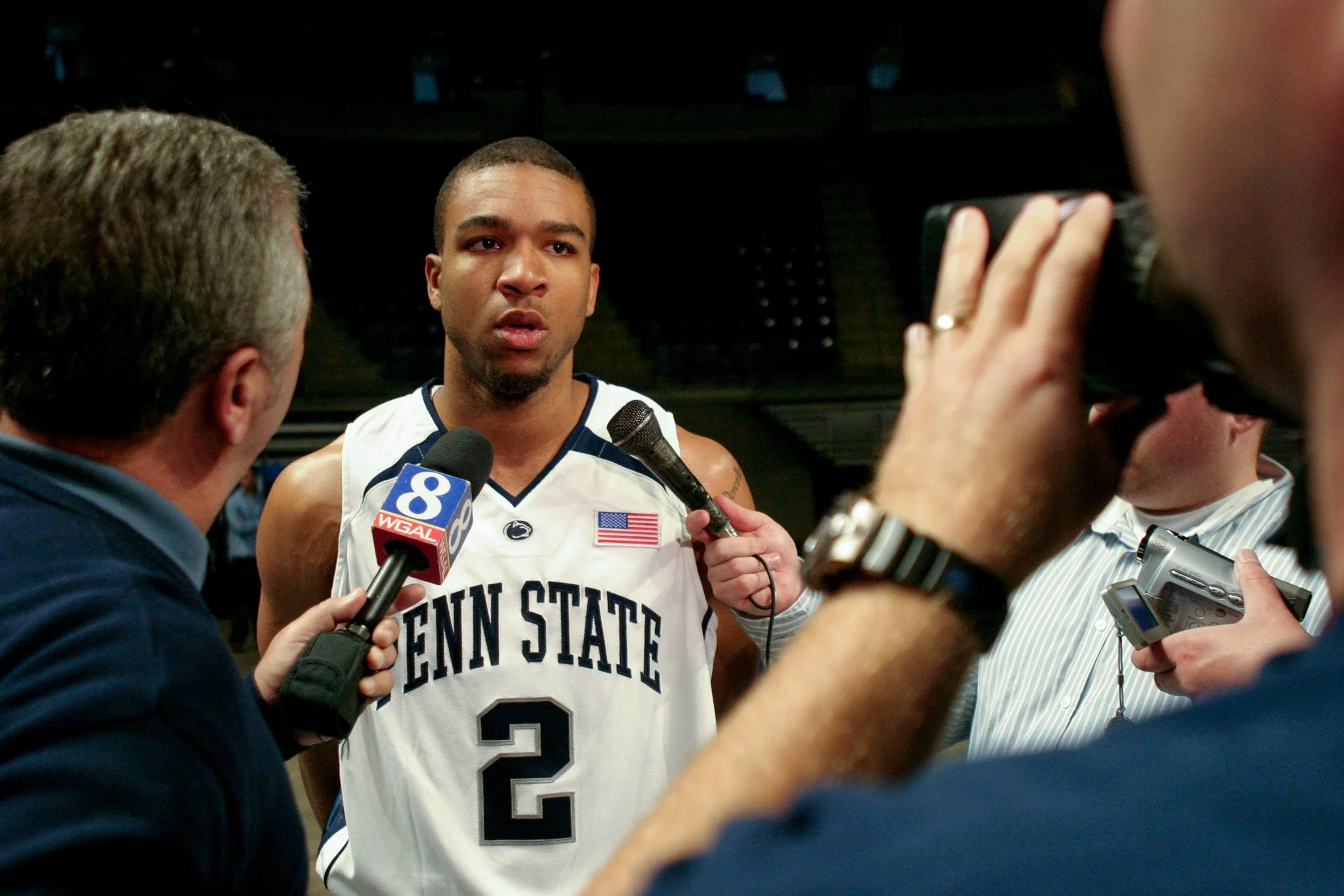 A basketball player from Penn State wearing a jersey with the number 2 is being interviewed by reporters. The player is surrounded by microphones and reporters, some holding recording devices. The background is dark and indoor.