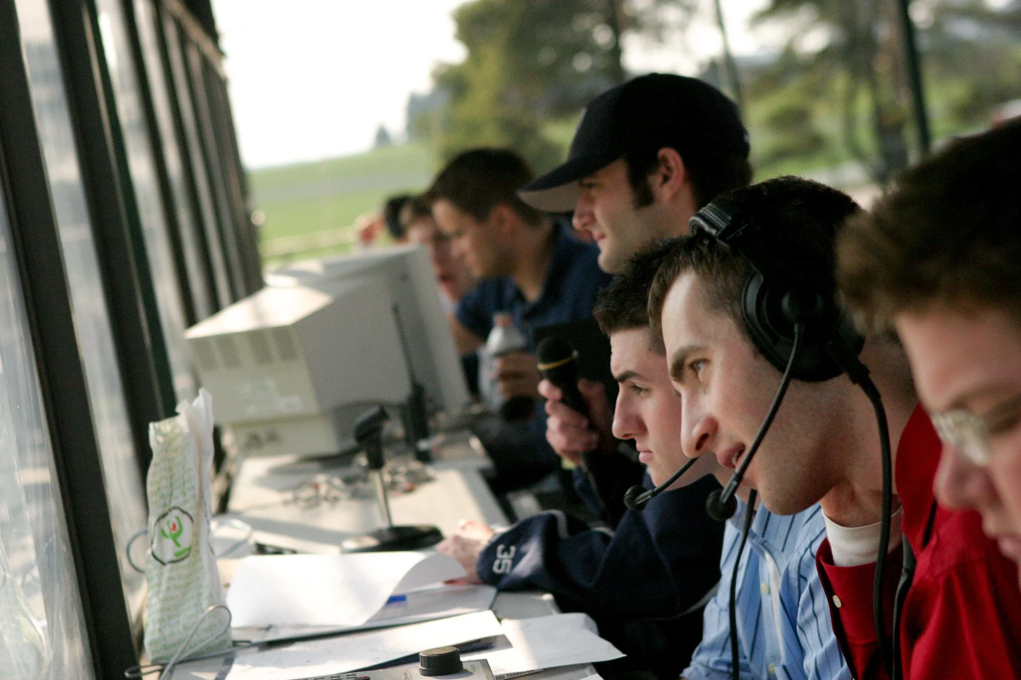 A group of young men working in a command center with multiple computer monitors, headsets, and papers, during daytime with green the outdoors visible through the window.