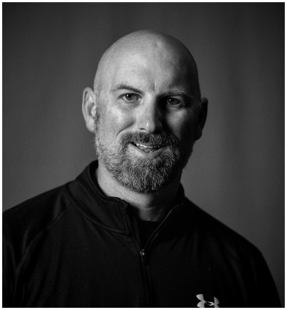 Black and white portrait of a bald man with a beard wearing a dark Under Armour jacket, smiling slightly against a plain background.