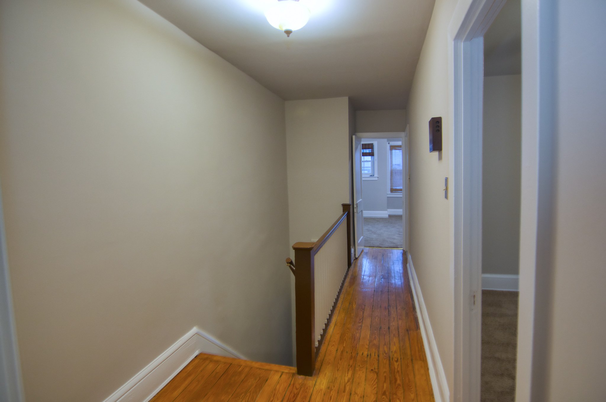 Empty hallway with wooden flooring, cream walls, and an open door leading to a brightly lit room with windows.