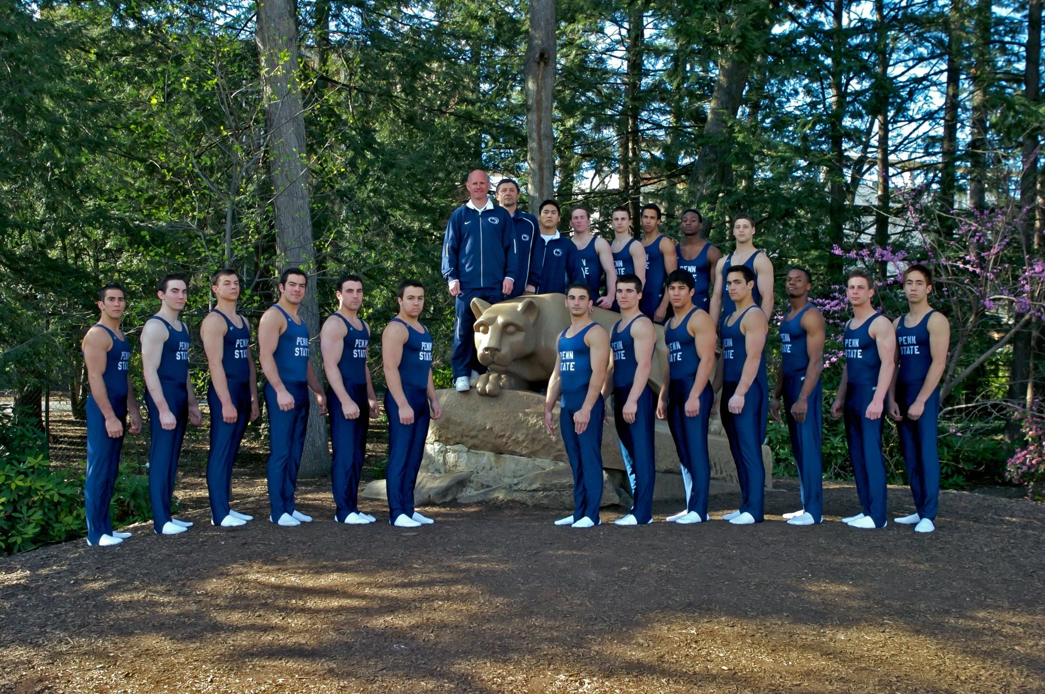 A group of male Penn State athletes in blue track suits standing outdoors around a sculpture of a lion's head in a wooded area.