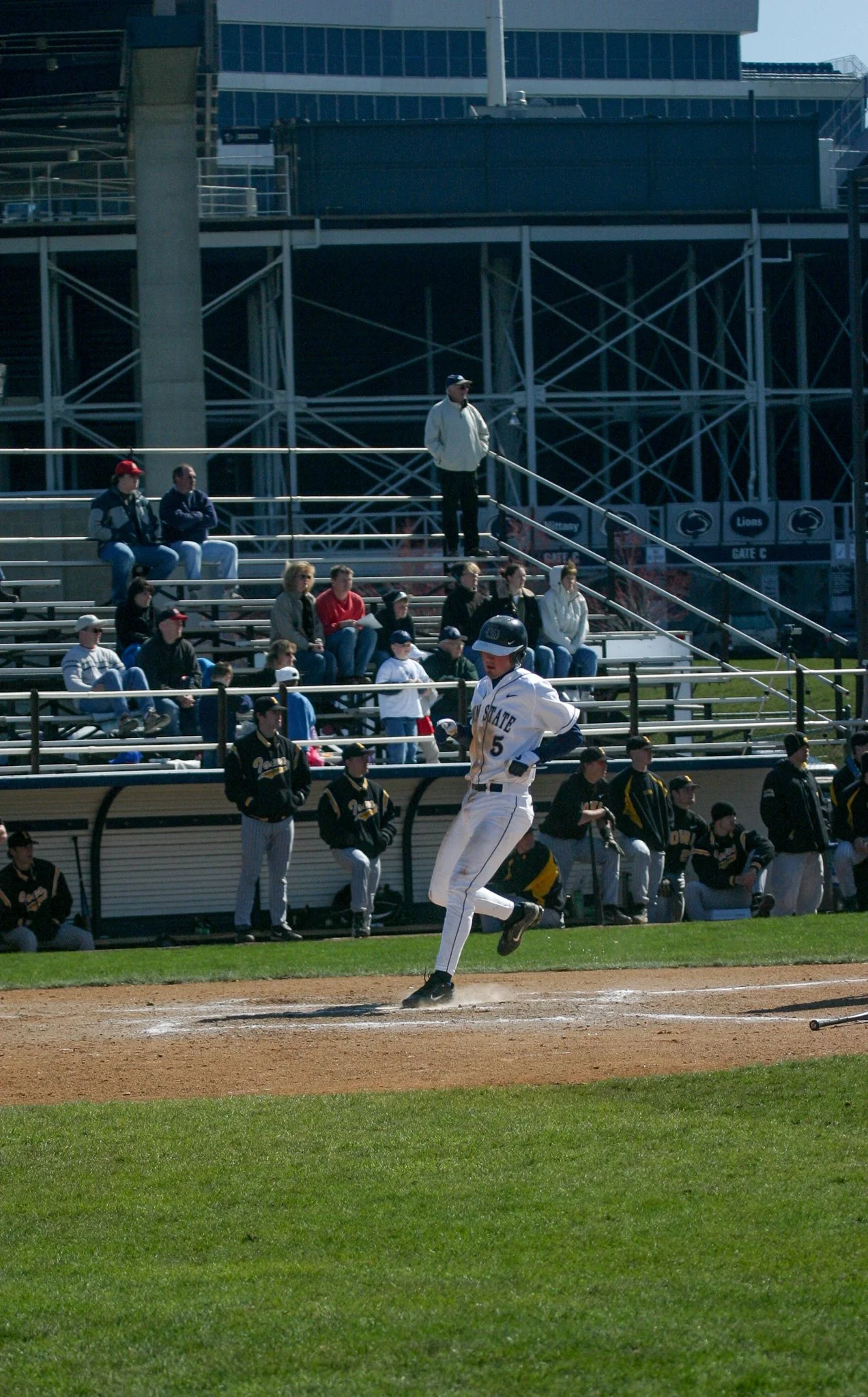 A baseball player running on the field during a game, with spectators sitting in the stands and other players on the bench in the background.