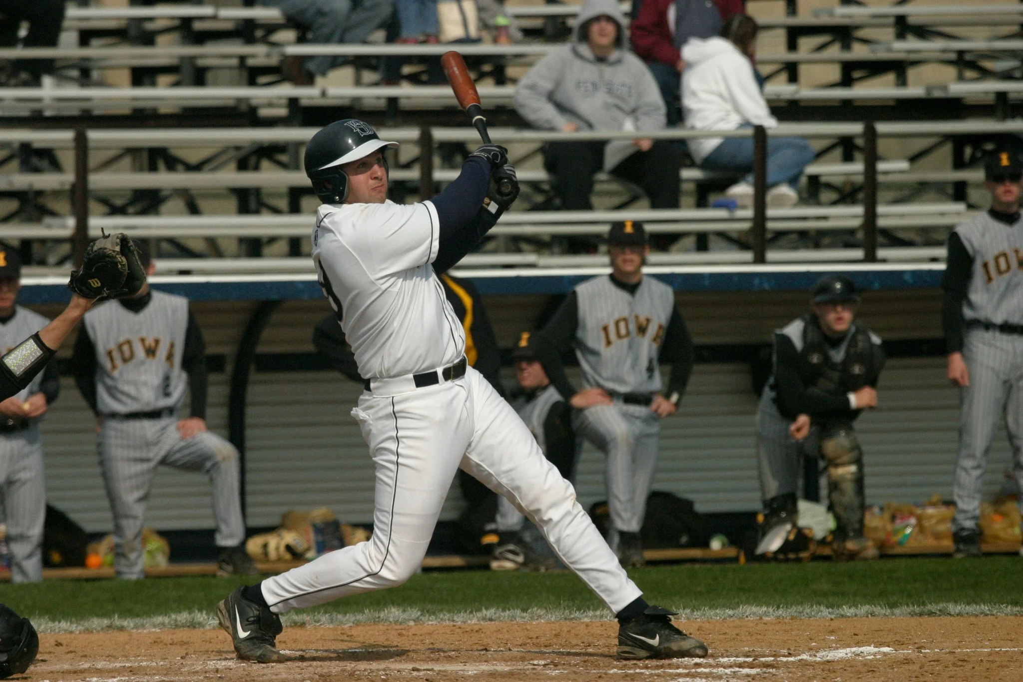 A baseball player in white uniform and black helmet swinging a bat during a game, with teammates and spectators in the background.